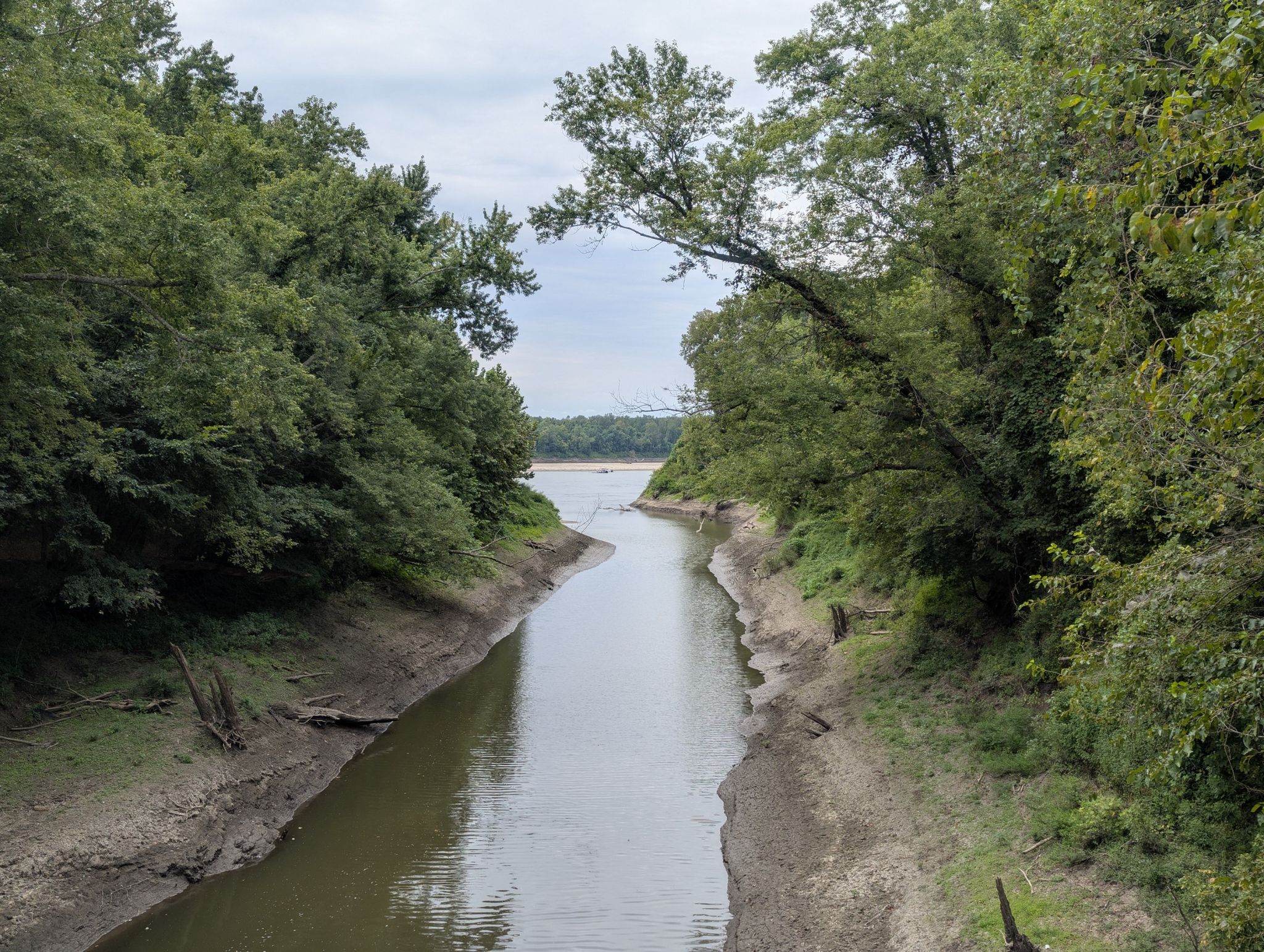 A narrow waterway flows between lush green trees under a cloudy sky. - Katy Trail: Hermann to Augusta