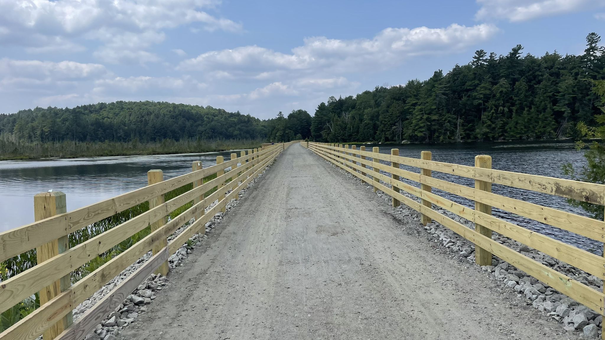 Long gravel path with wooden fences, flanked by water and distant forests. - Lake Flower – Colby Lake Bridge loop from Lake Clear