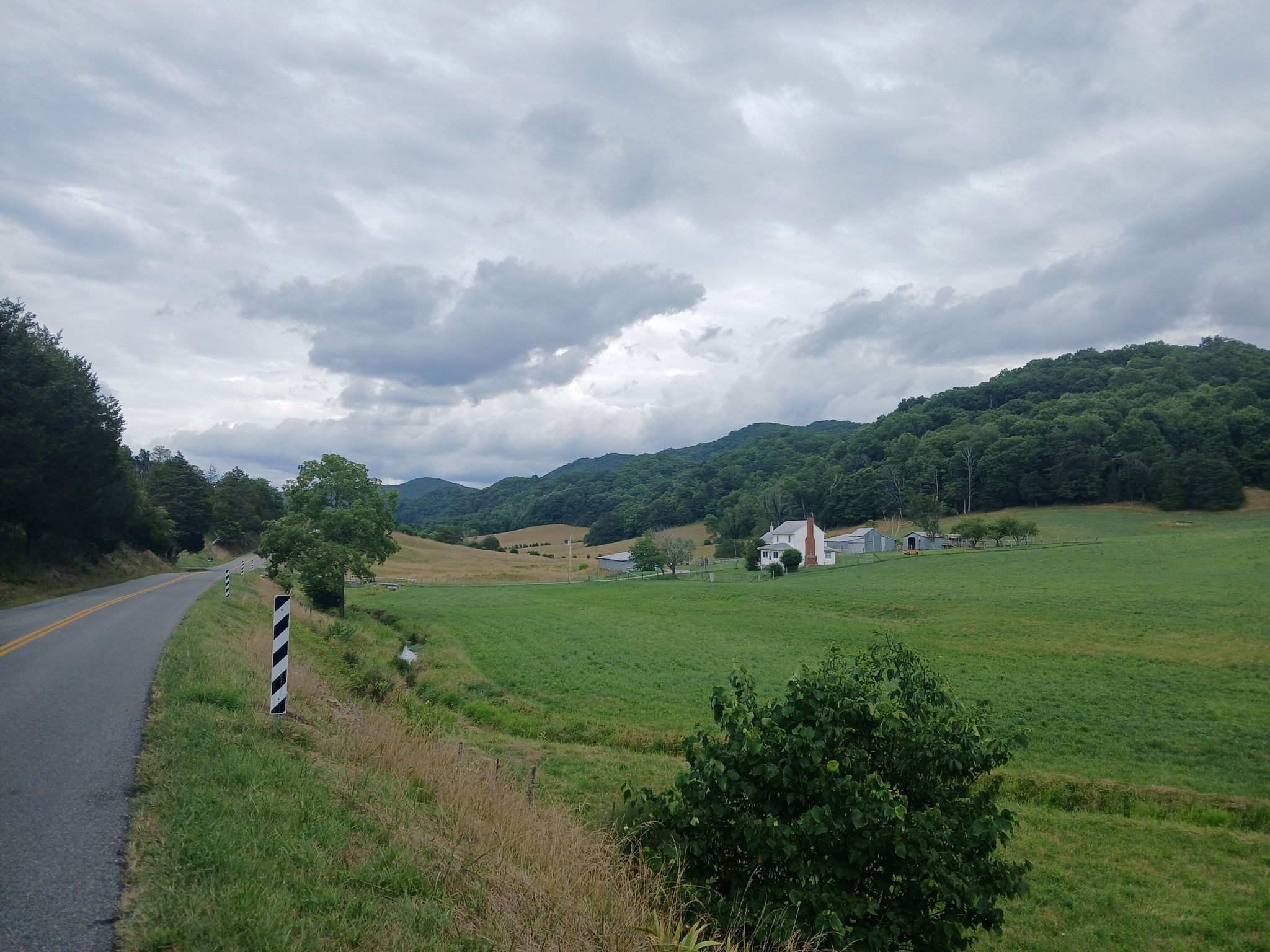Paved road, green fields, white house, and forested hills under cloudy skies. - Willow Springs Lake loop