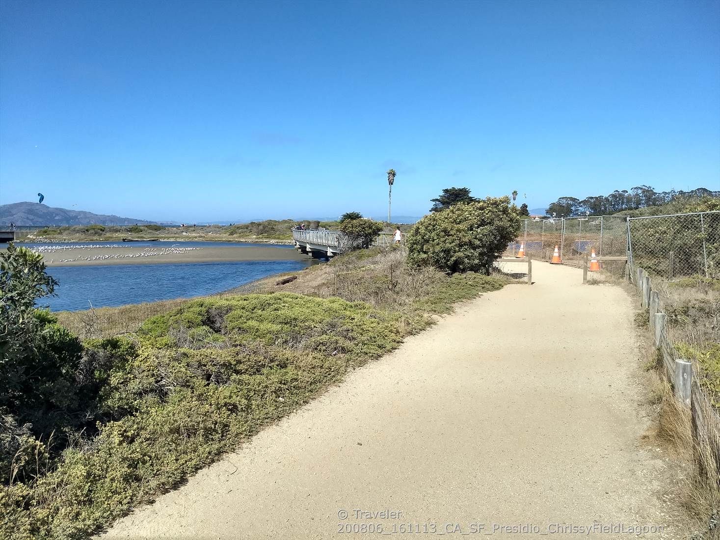 A dirt path curves beside a calm body of water under a clear sky. - San Francisco Bay Trail