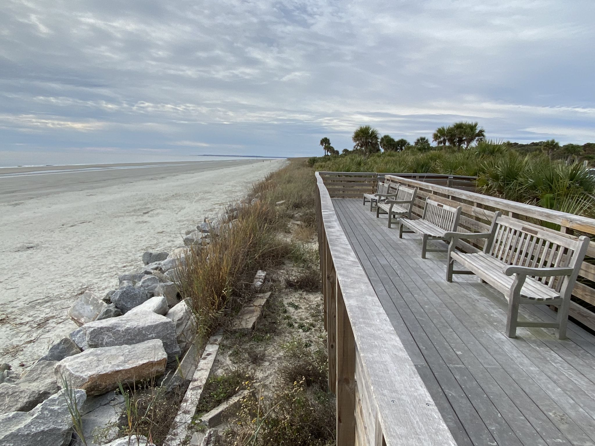 A wooden boardwalk with benches overlooks a wide sandy beach under a cloudy sky. - Jekyll Island Beach Park loop from Tidelands Nature Center