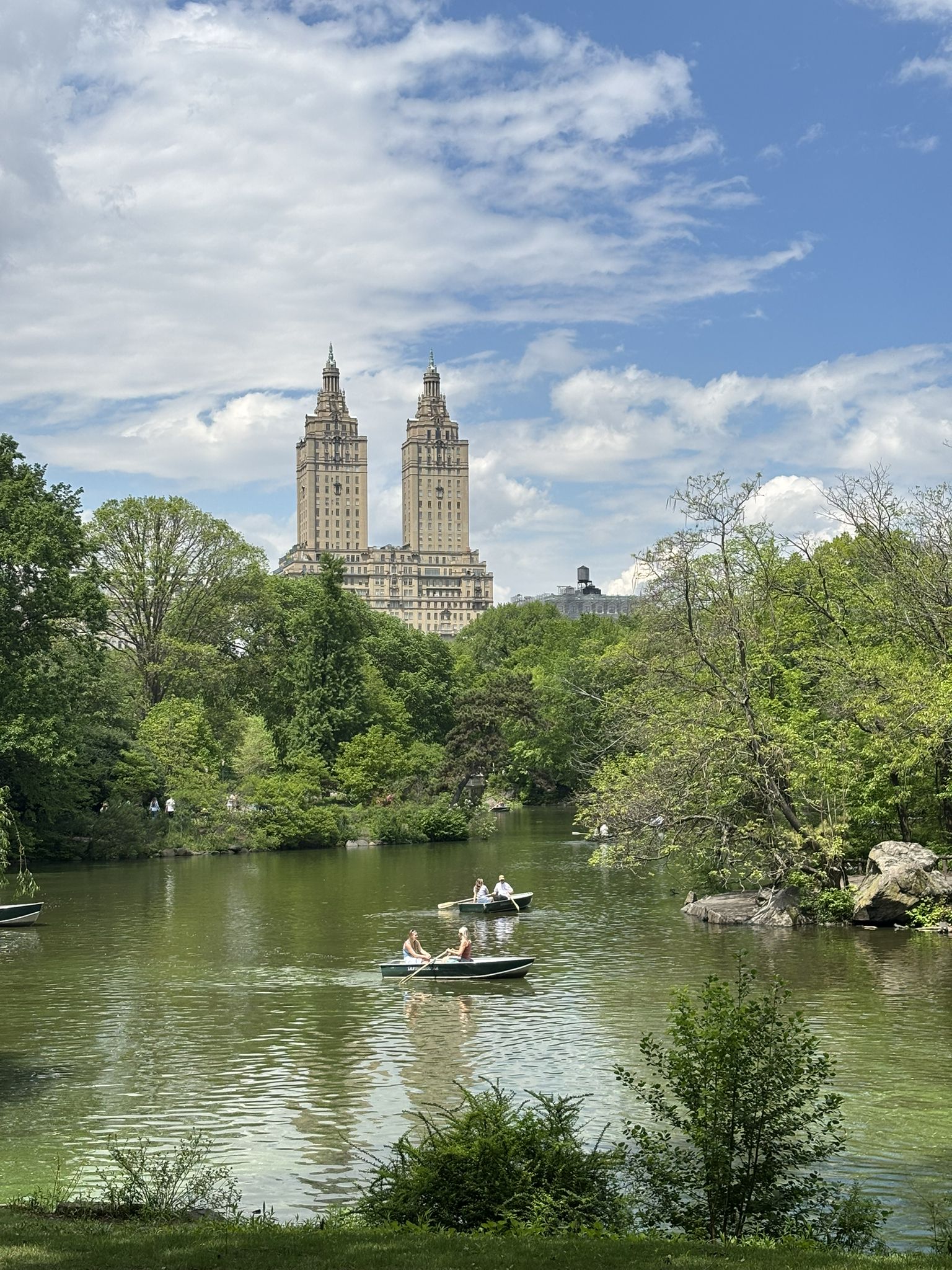 Lake with rowboats, prominent twin buildings, green trees, partly cloudy sky. - View of the San Remo Towers – Bethesda Fountain and Terrace loop from 59th Street