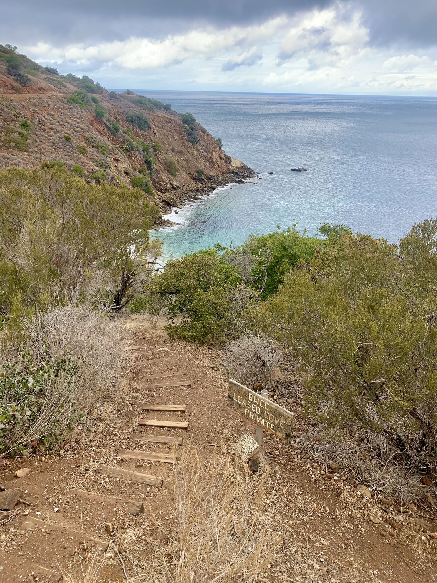 Dirt path descends to a rocky cove with turquoise water under an overcast sky. - View of Big Geiger Cove – View of Cherry Cove loop from Indian Rock