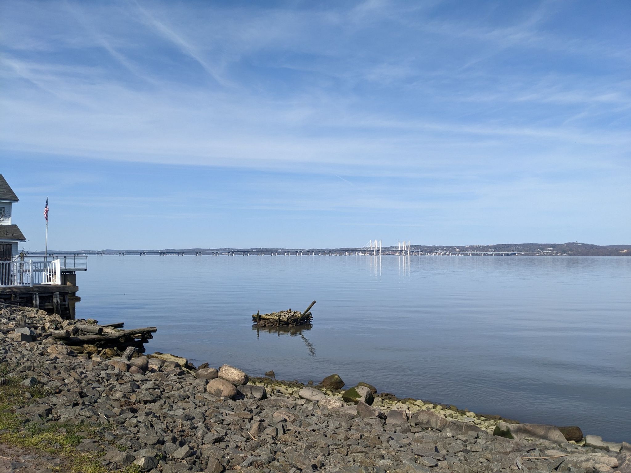 Calm water with a distant bridge, rocky shore, and a house on a clear day. - View of the Hudson River – Piermont Railway Station (historic) loop from Englewood