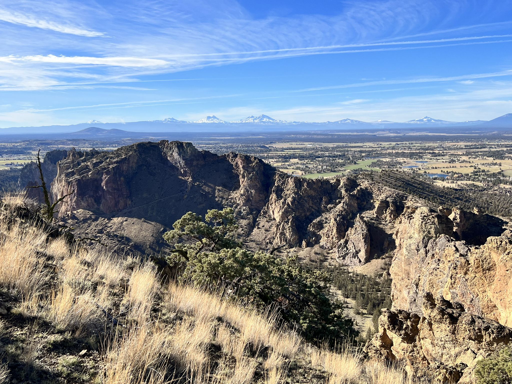 Rugged cliffs overlook a vast valley with distant snow-capped mountains under a blue sky. - Gray Butte Trail