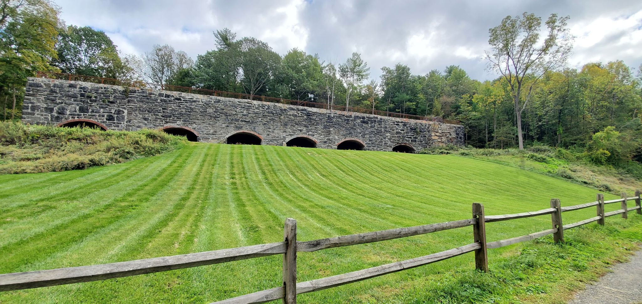 A stone arch bridge overlooks a manicured green grassy slope with a wooden fence. - Cold Air Cave – Old Cementworks loop from Rosendale