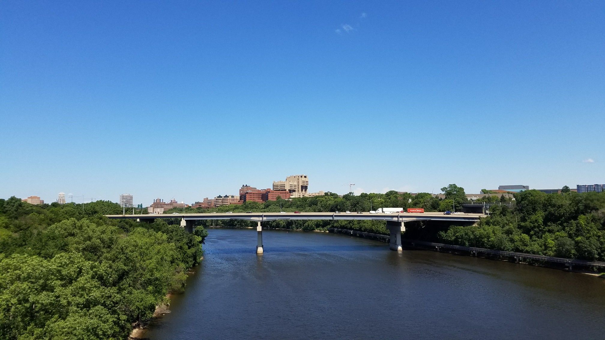 Wide river flowing under a long bridge, surrounded by green trees. - Martin Olav Sabo Bridge – Cedar Lake loop from Westgate