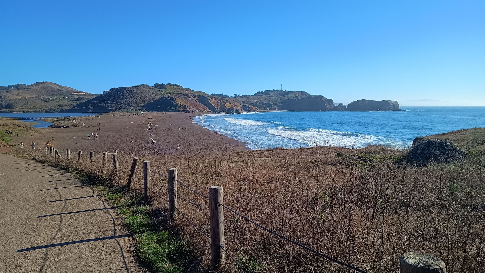 Sunny beach with ocean waves, coastal hills, and people walking on sand. - Rodeo Valley Trail – Battery Townsley loop from Golden Gate