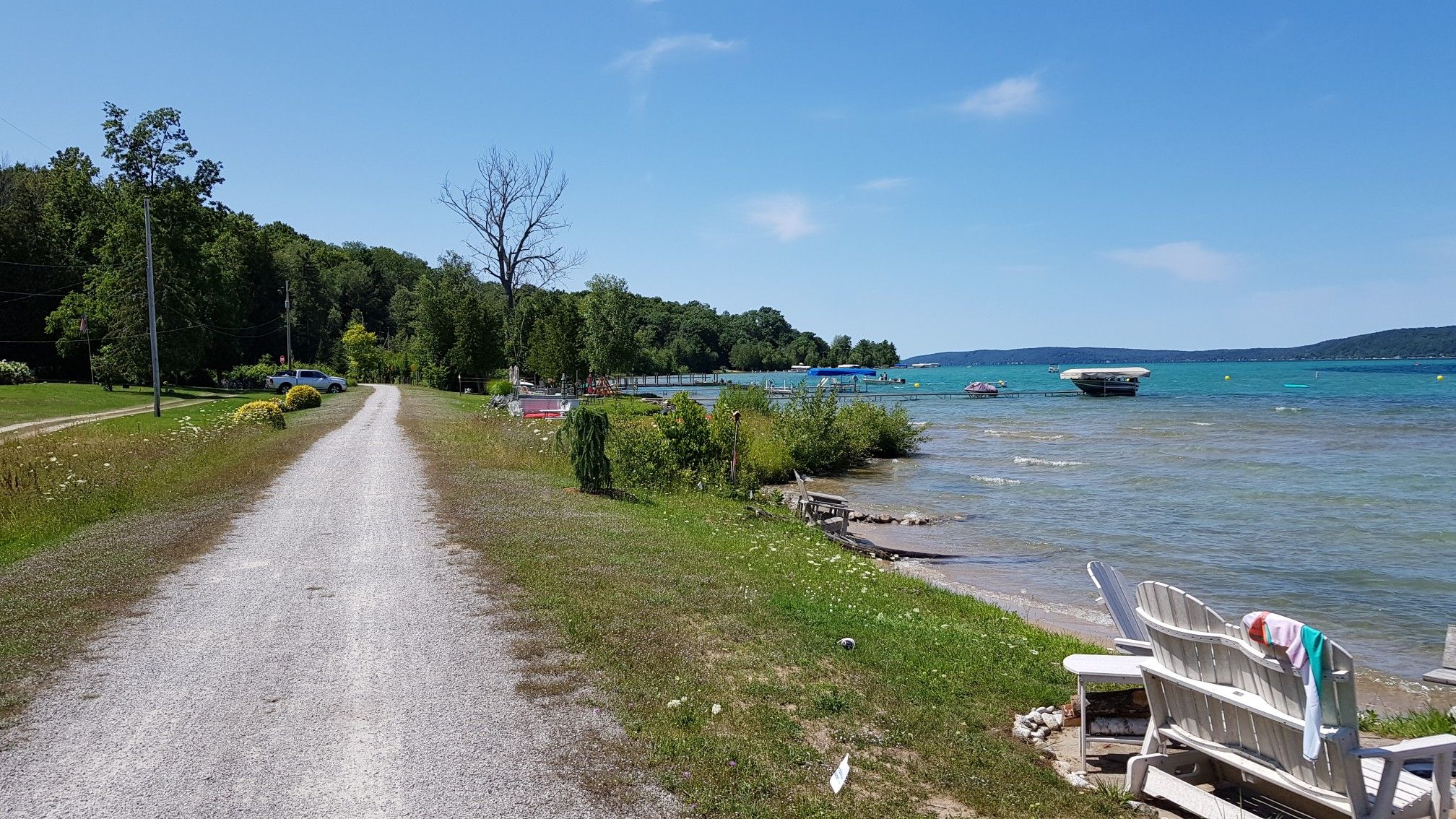 Gravel path beside a blue lake with white chairs and distant hills on a sunny day. - Beulah Beach loop from Benzonia Township