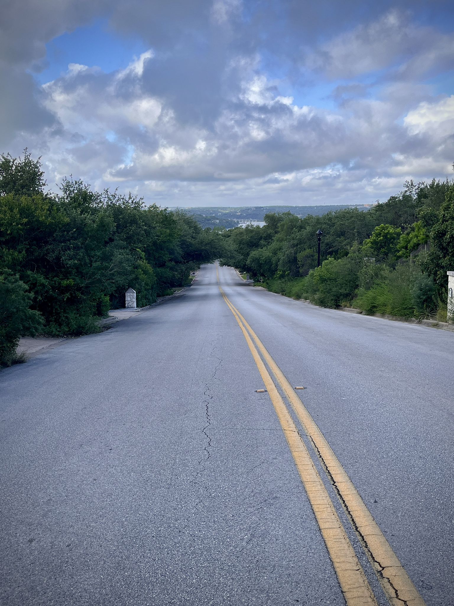 Empty road descends through green trees under a cloudy sky towards distant water. - River Place Nature Reserve & Lake Austin loop