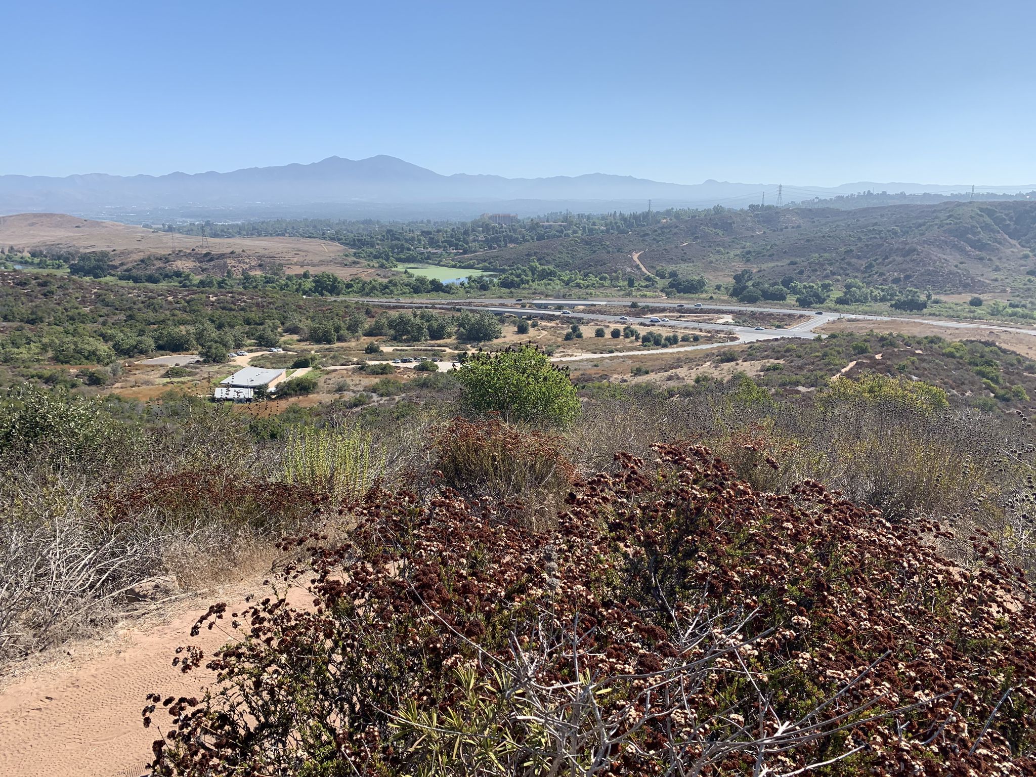 Overlook of a wide valley with dry brush, distant mountains, and a building. - Little Sycamore Canyon and Serrano Ridge Loop