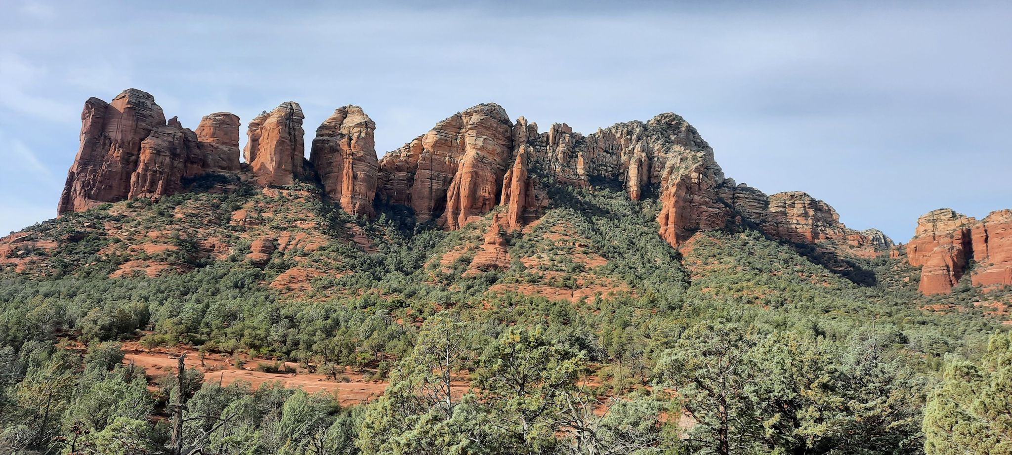 Red rock formations rise above a green forested hillside under a partly cloudy sky. - Soldier Pass Trail
