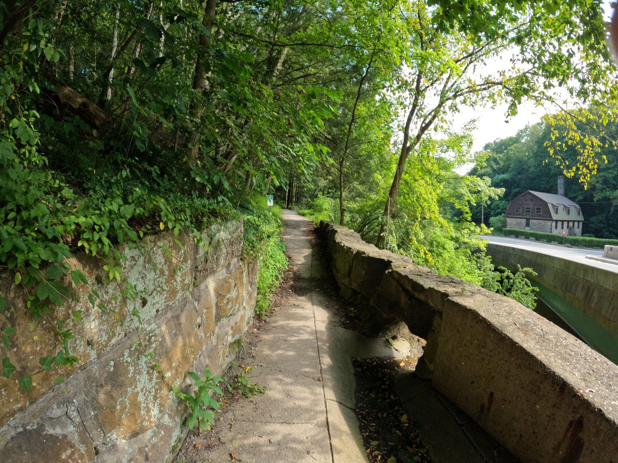 A concrete path next to a stone wall, with green trees and a building ahead. - View of Lanterman Falls – Lantermann Falls Covered Brigde loop from Mill Creek Park