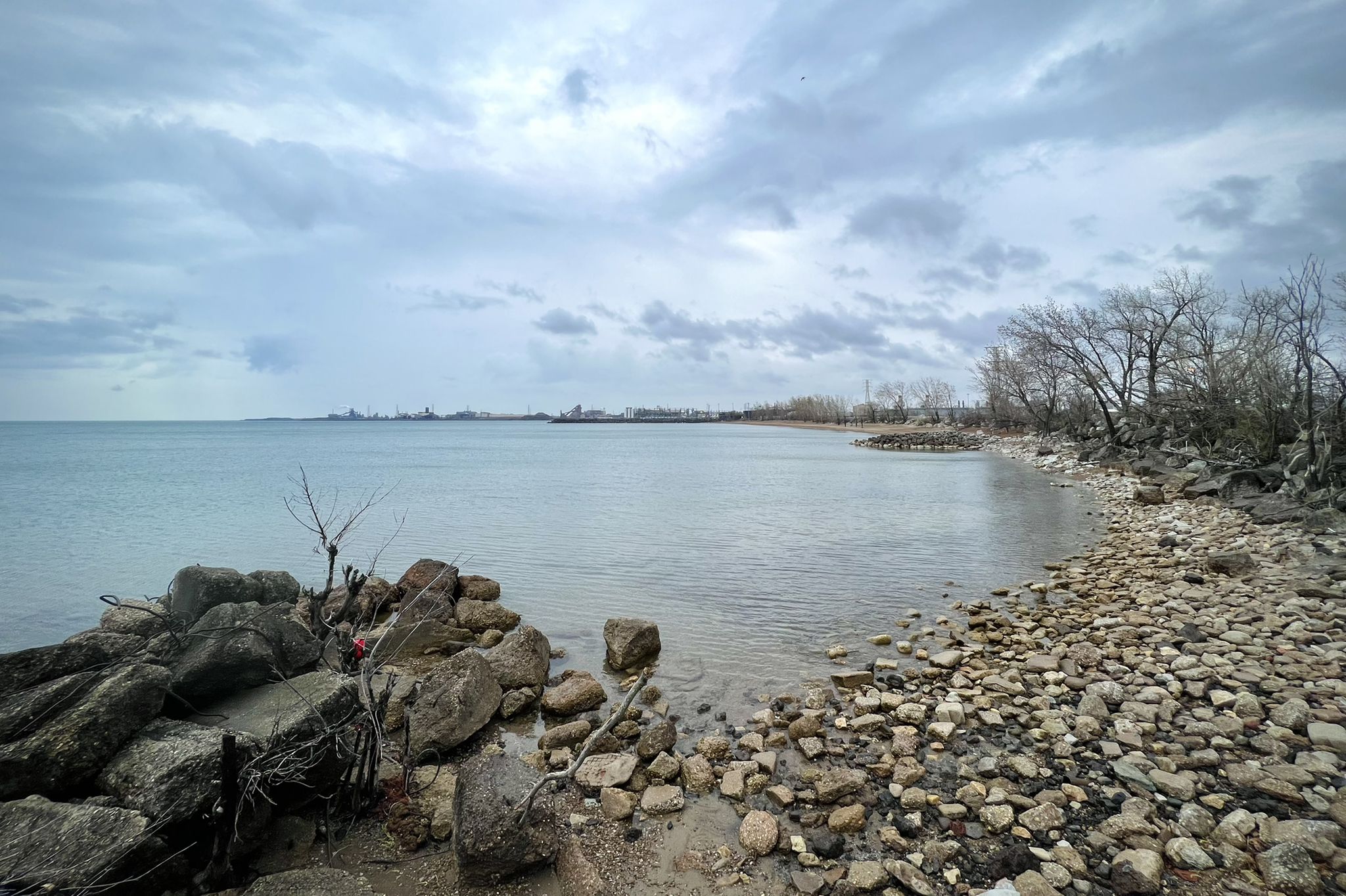 Overcast day on a rocky shoreline with calm water and distant city. - Wolf Lake and Whihala Beach Loop