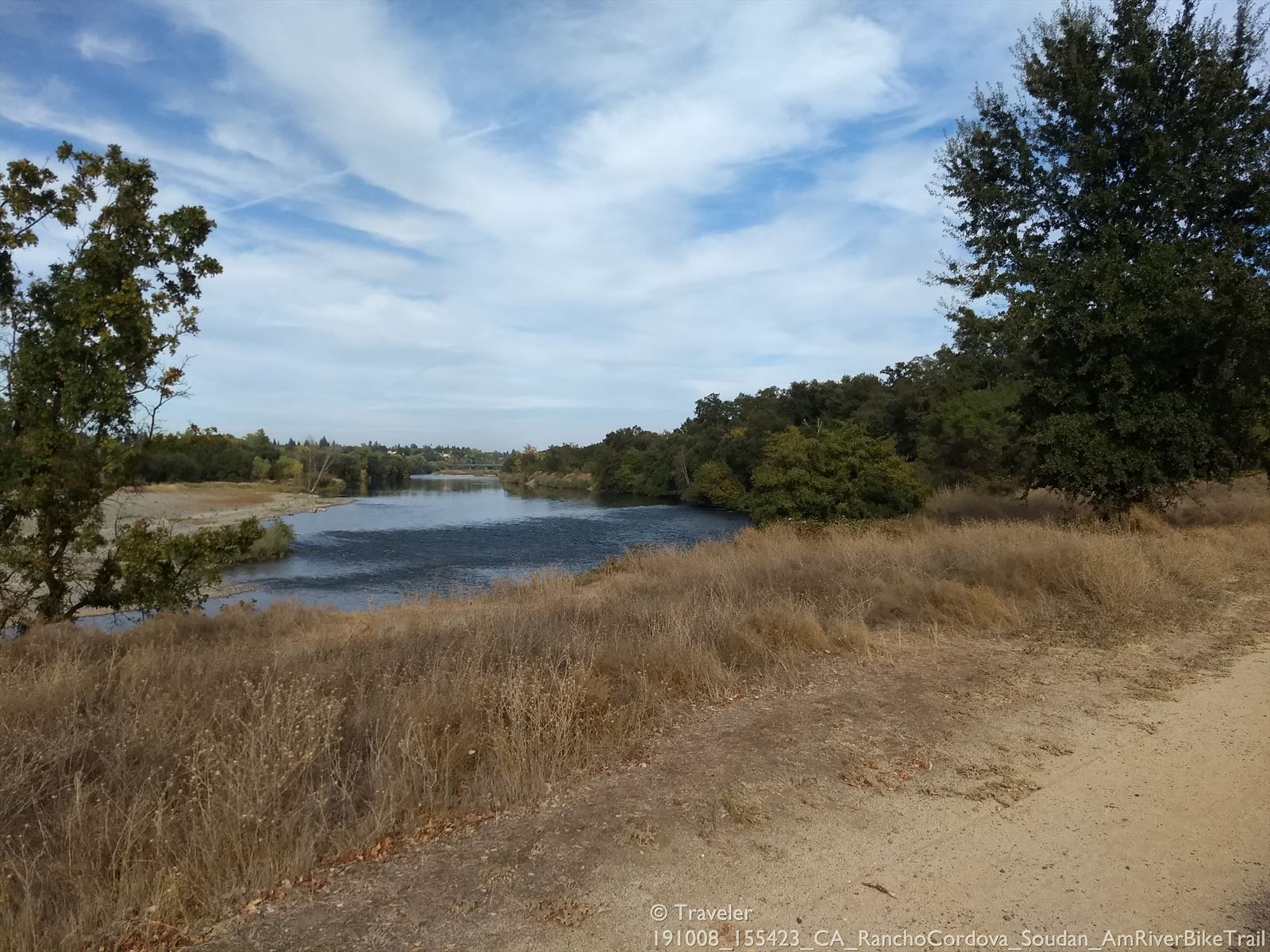 River flowing past dry grass and dense green trees under cloudy sky. - American River Bike Trail