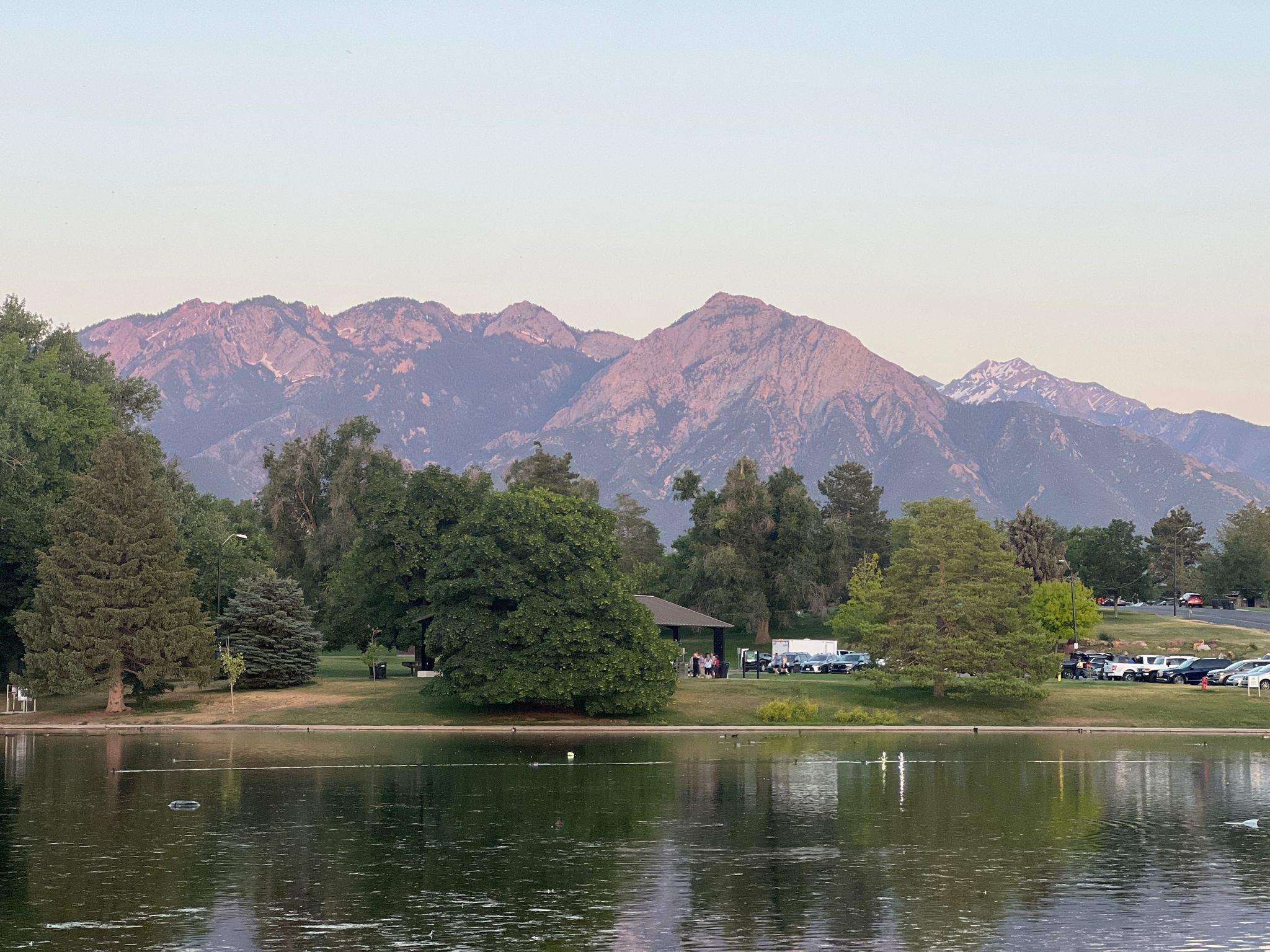 A park lake with trees and distant mountains under soft, warm light. - Sugar House Park Loop