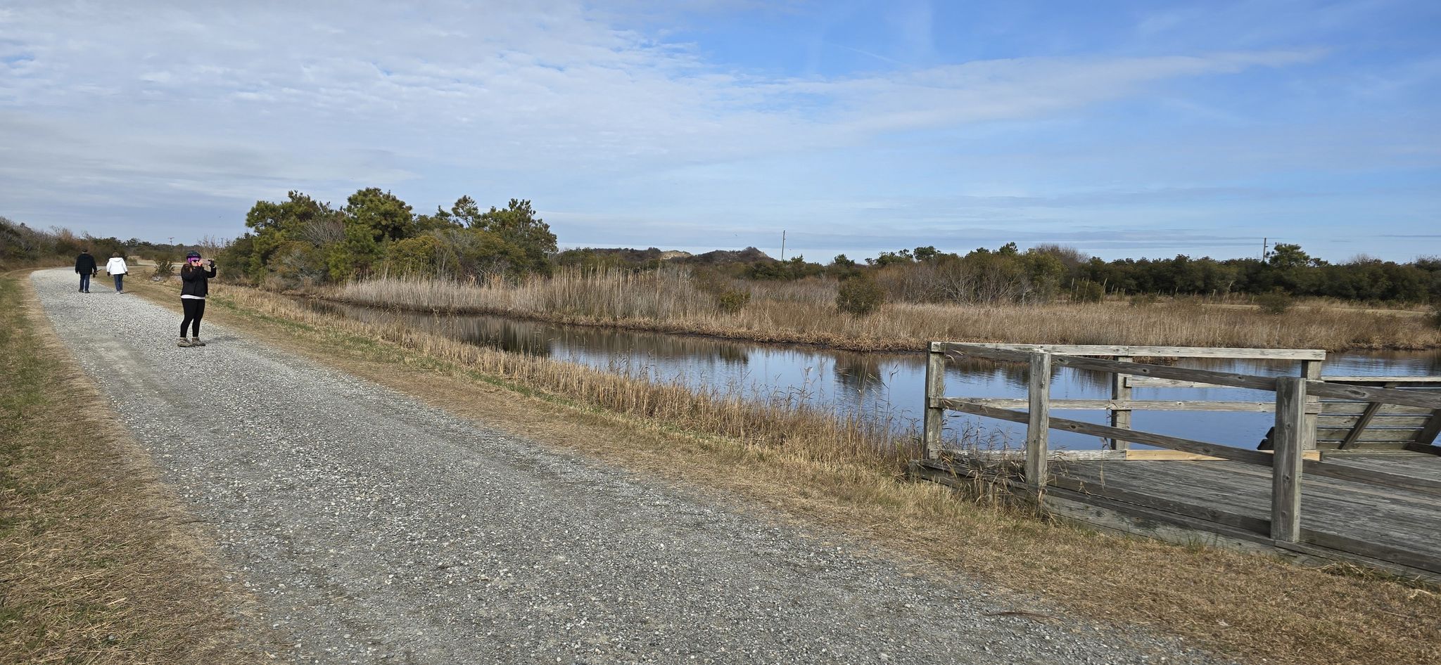 People walk on a sunny gravel path beside a calm reedy waterway. - Dunes and Seaside Trail