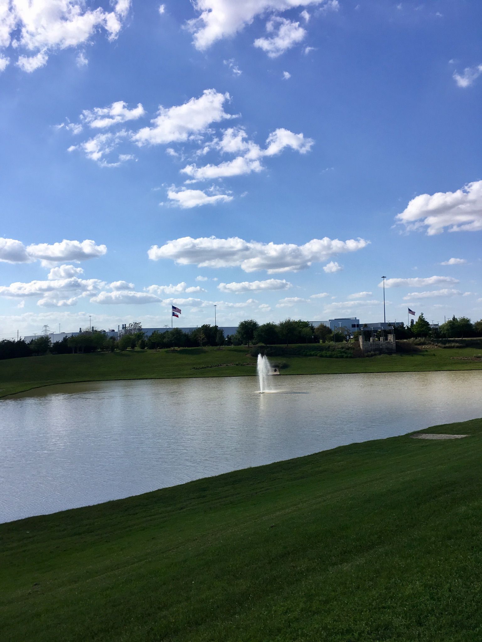 A lake with a fountain, green grass, and distant buildings under a cloudy sky. - Rick Rice Park loop from James E Taylor High School