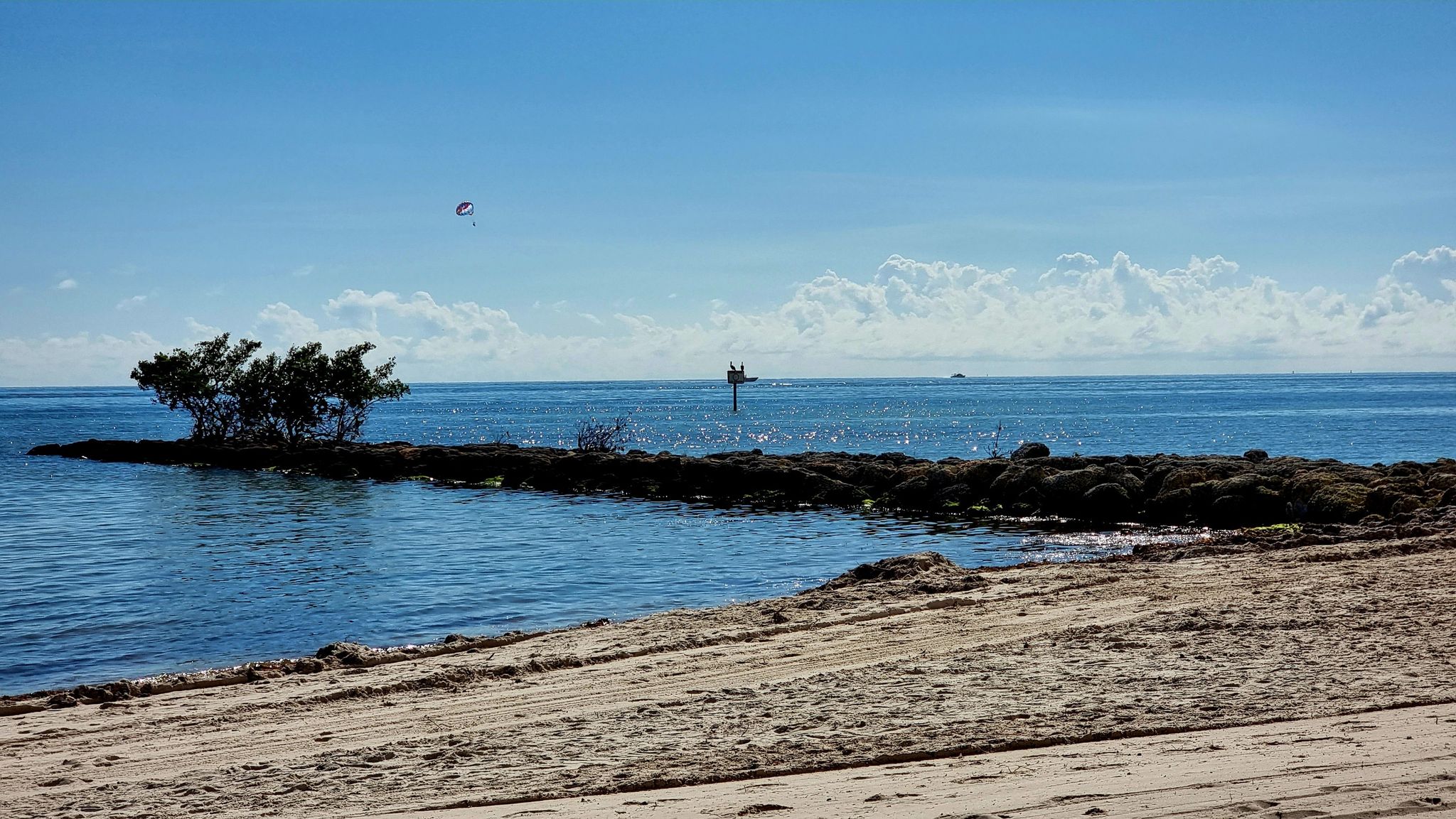 Calm ocean with a small tree-covered island and distant kite under blue sky. - Southernmost Point Buoy – Mile 0 of U.S. Highway 1 loop from Key West