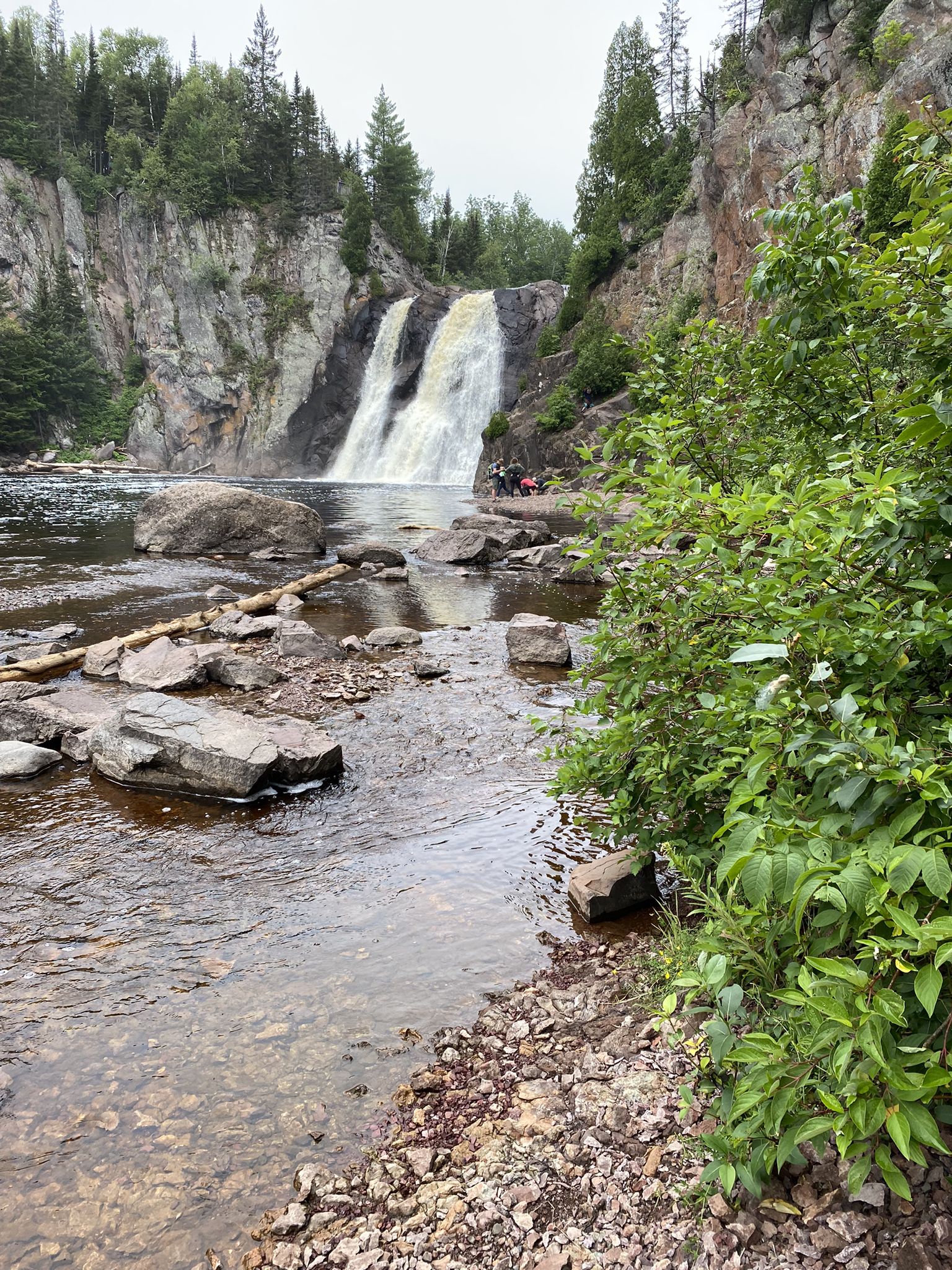 Two waterfalls flowing into a rocky river, with green foliage and distant people. - High Falls and Two Step Falls Loop