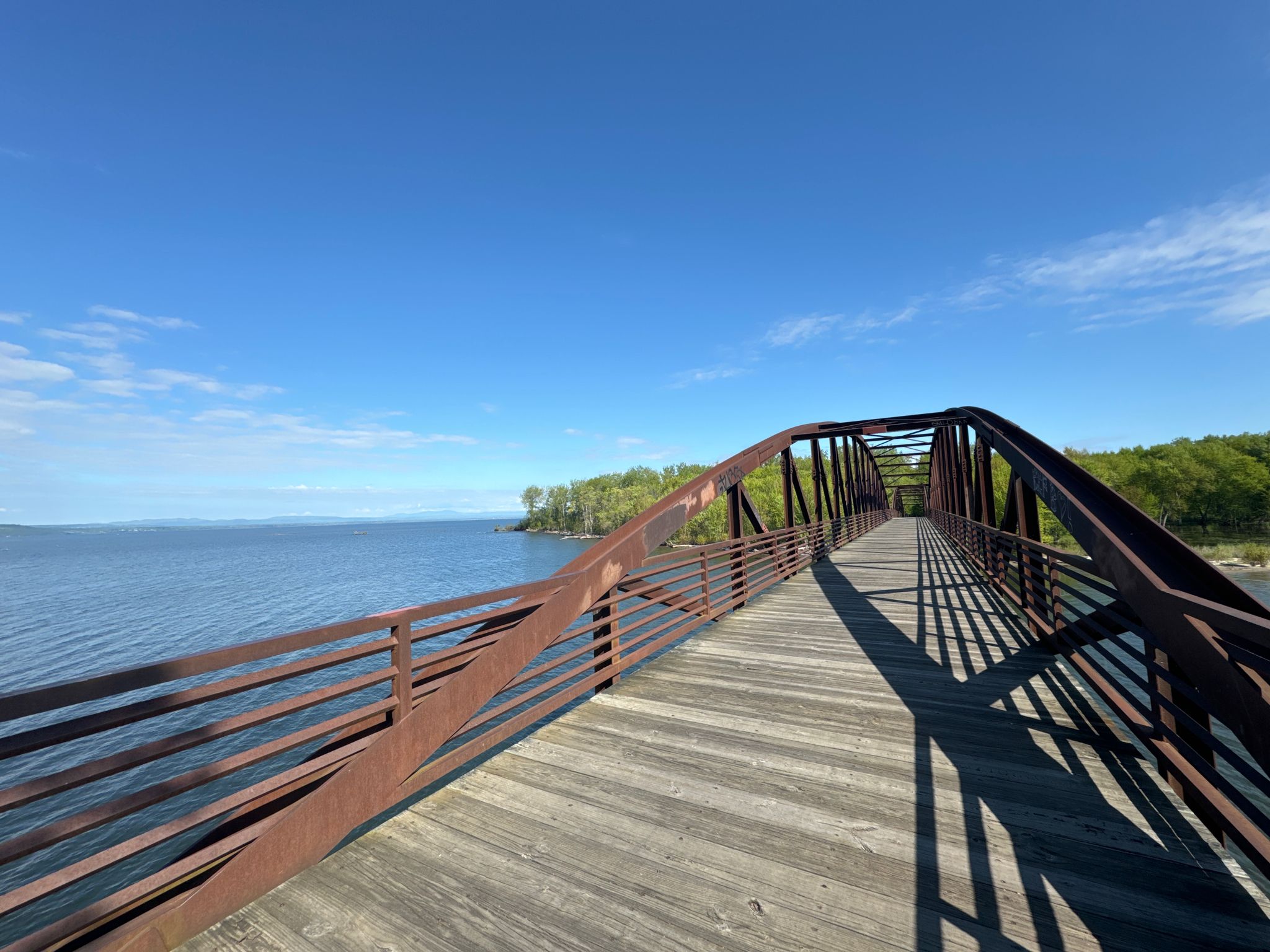 A long, rusty metal bridge extends over calm blue water under a clear sky. - Granite Steps – Colchester/Burlington bridge loop from Burlington