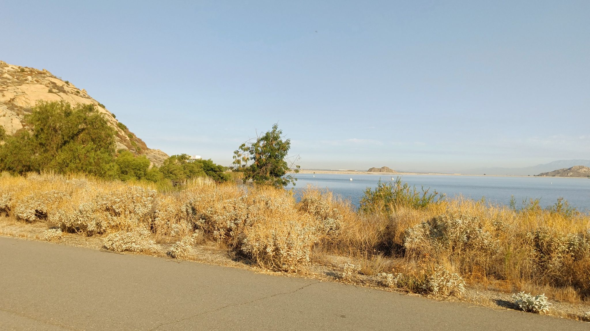 Paved road alongside dry bushes overlooking a vast lake and hills. - Lake Perris – Scenic Cycling Spot loop from Lake Perris