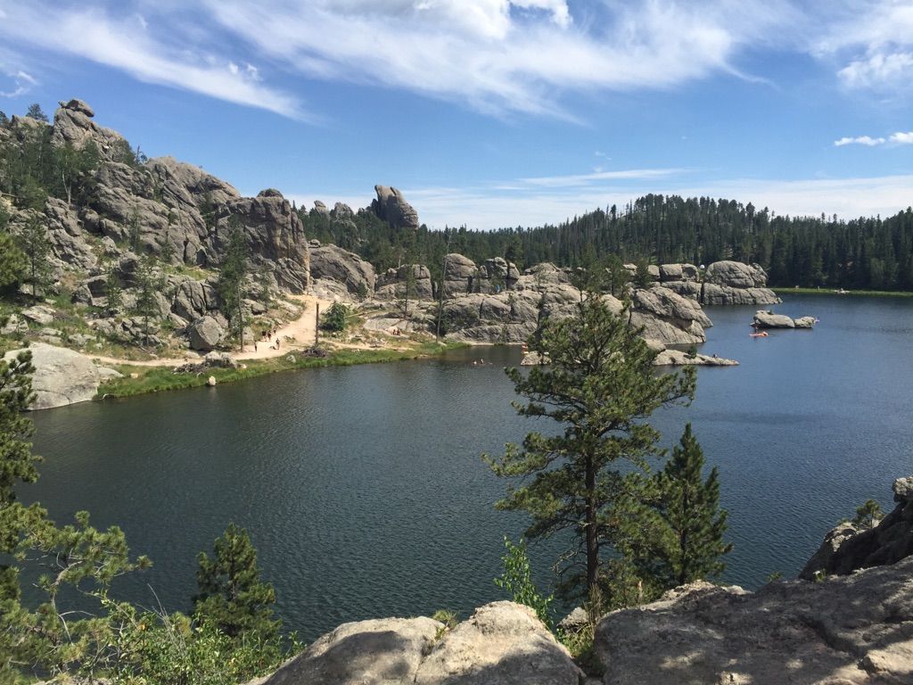 Lake with rocky shores and pine trees under a partly cloudy sky. - Sylvan Lake Shore Trail