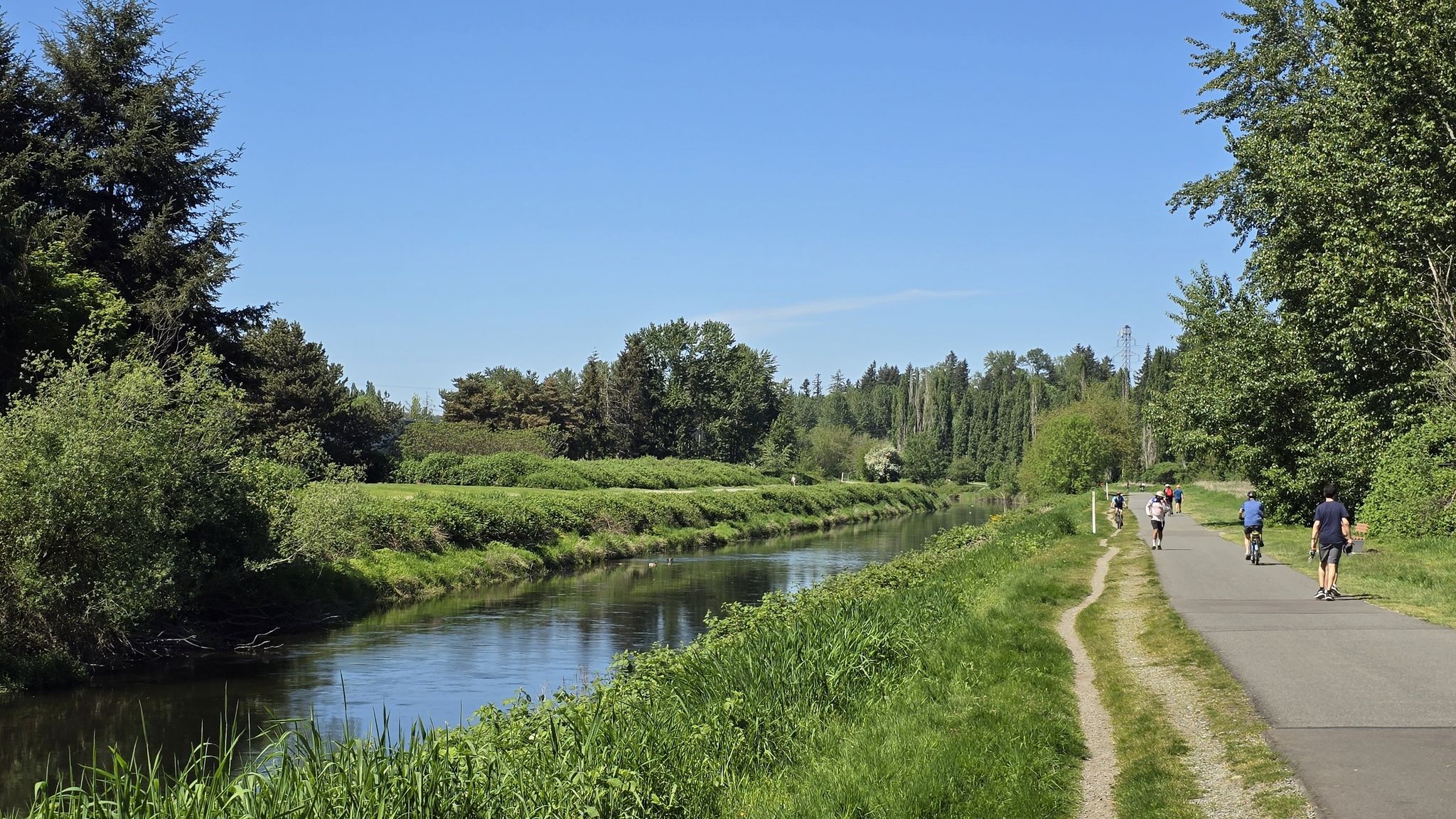Sunny day, people walk and cycle on a paved path beside a calm river. - Lake Washington Loop