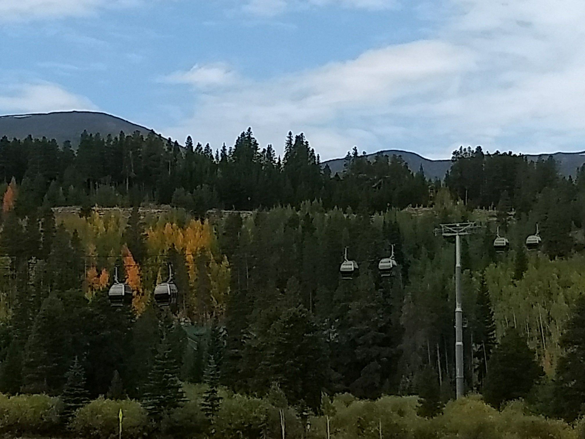 Gondolas ascend a forested mountain with autumn colors under a partly cloudy sky. - Upper Blue Recpath