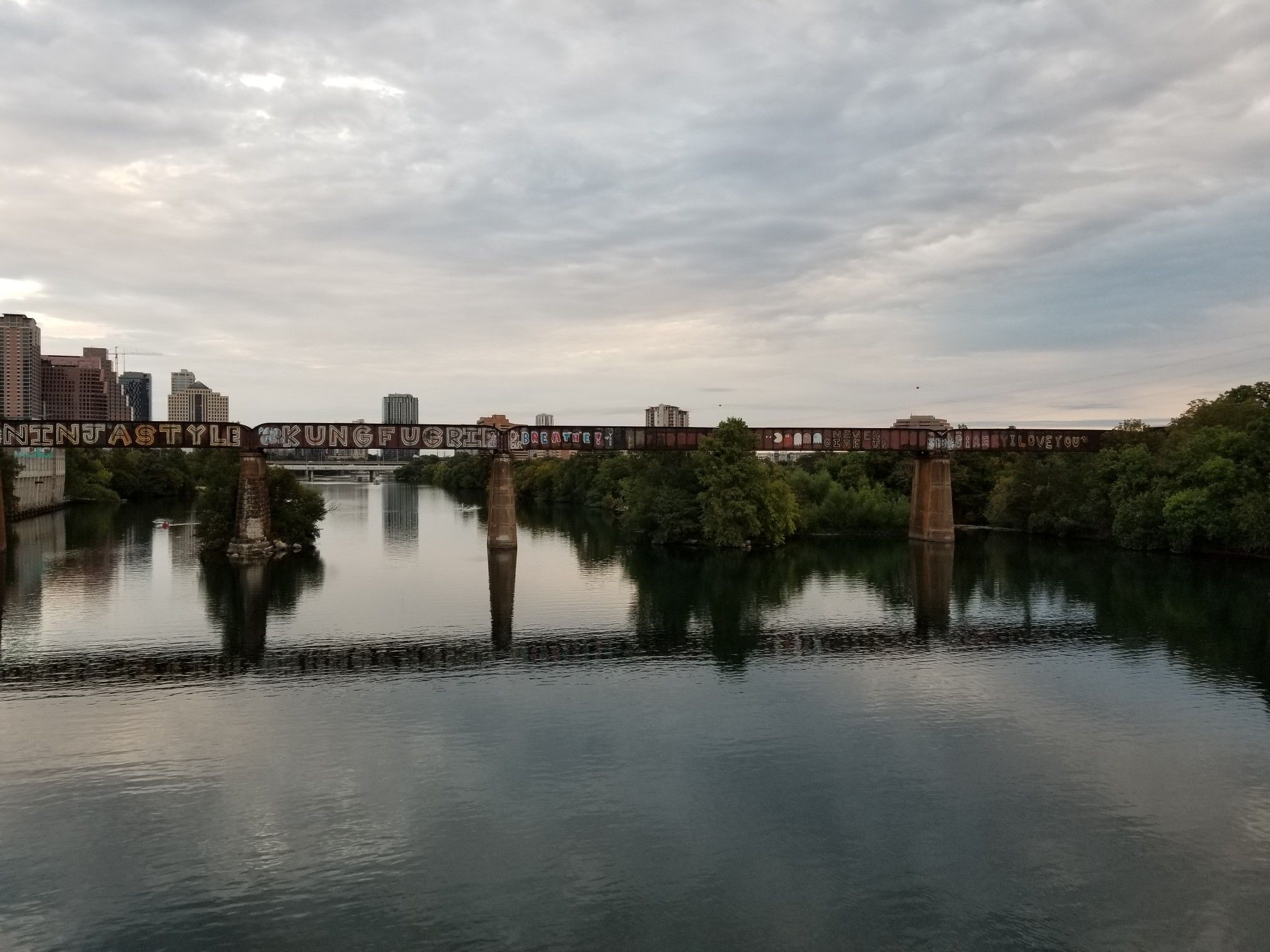Overcast view of a wide river, a long train bridge, and city buildings. - Red Bud Isle – Pfluger Pedestrian Bridge loop from Barrow Nature Preserve