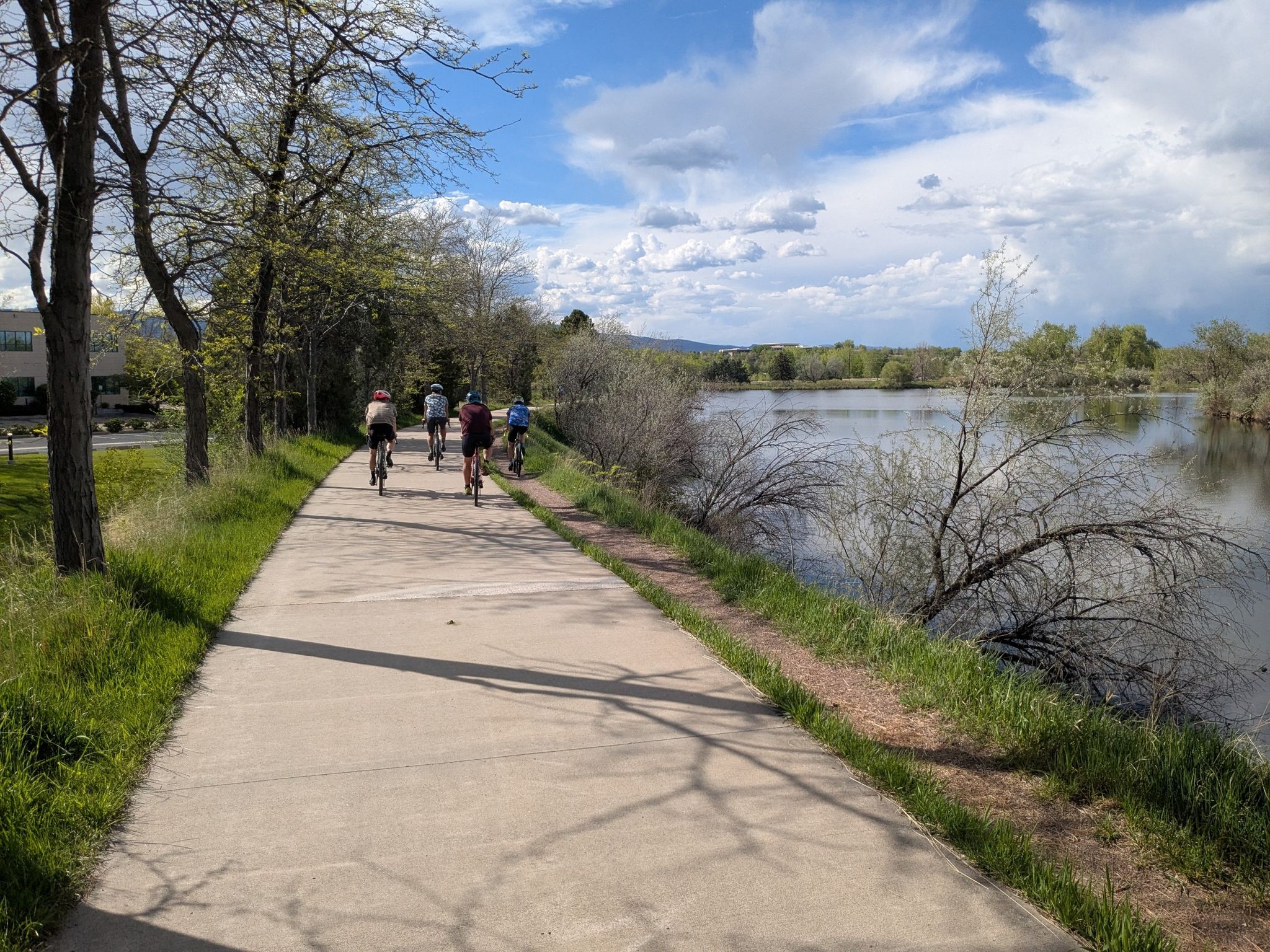 Cyclists ride a paved path beside a calm lake on a bright day. - Coot Lake Dock – Twin Lakes loop from Valmont