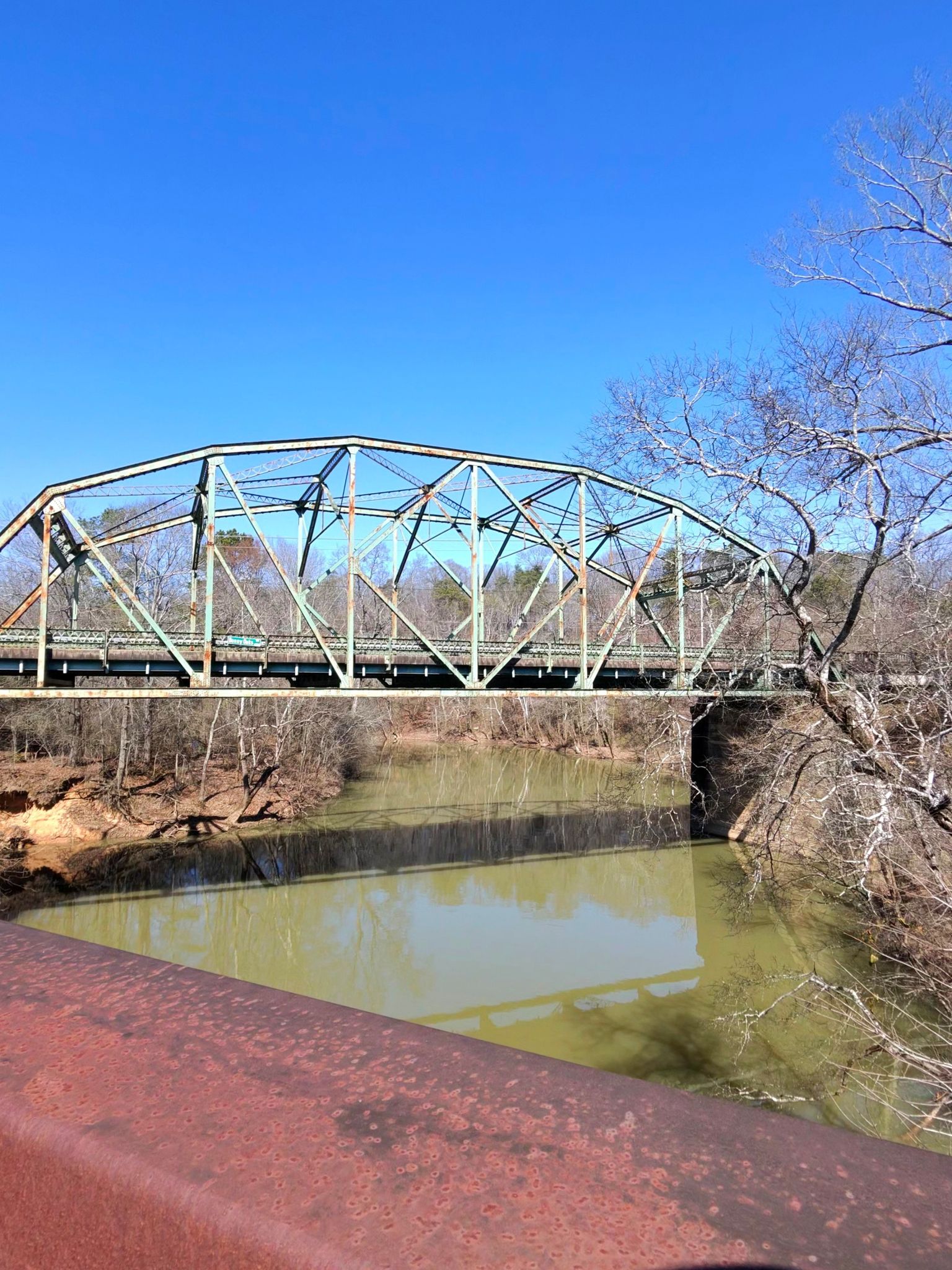 A green metal truss bridge spans a calm river under a clear blue sky. - South Chickamauga Creek Greenway