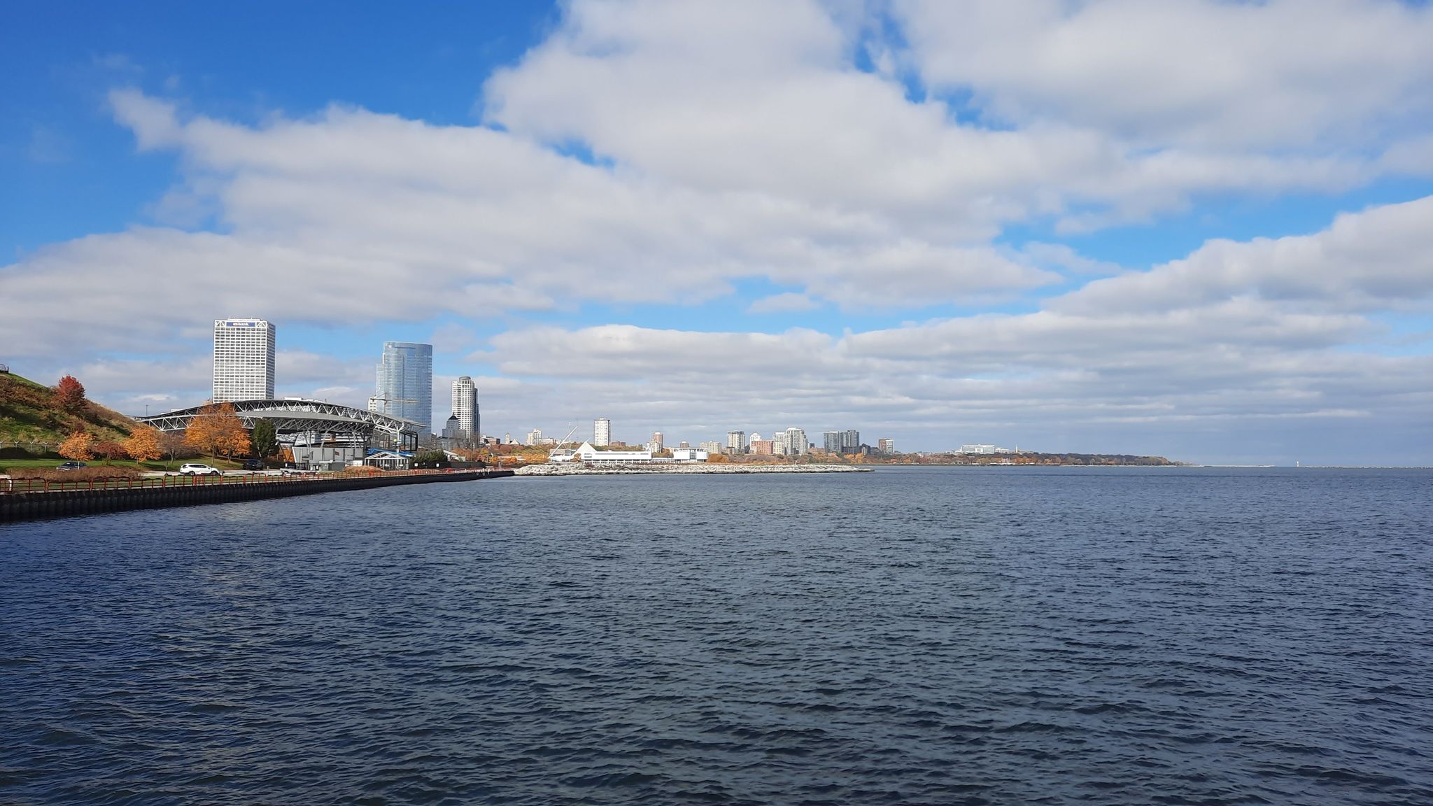City skyline across a wide body of water under a partly cloudy sky. - Milwaukee Pierhead Lighthouse via North River Walkway