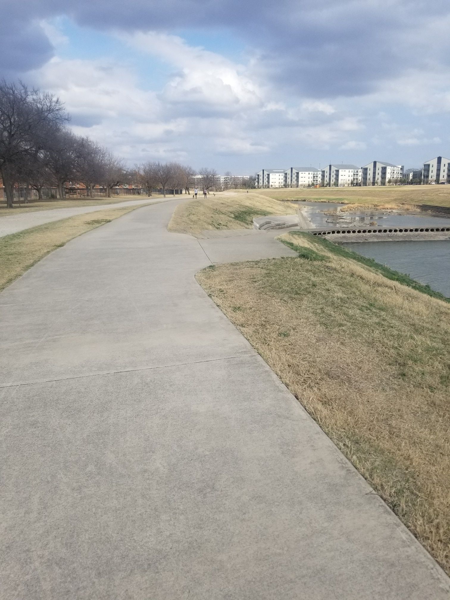 Paved path next to a river with dry grass and distant buildings. - Clear Fork Trinity River Trail – Benbrook Lake loop from Ridglea Country Club Parking