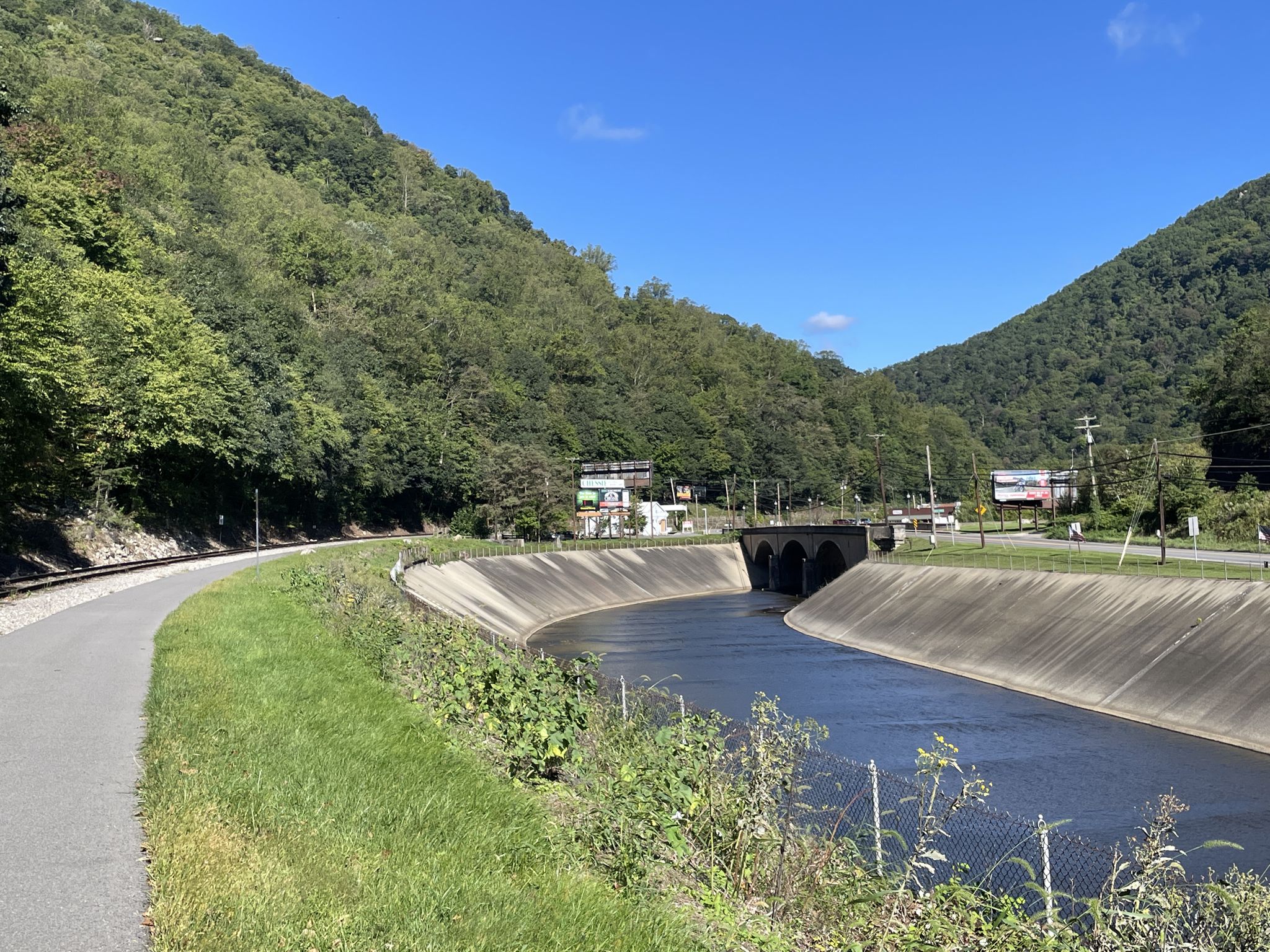 Concrete canal, paved path, and railroad tracks alongside green, tree-covered mountains. - Helmstetter’s Curve – Brush Tunnel loop from Cumberland