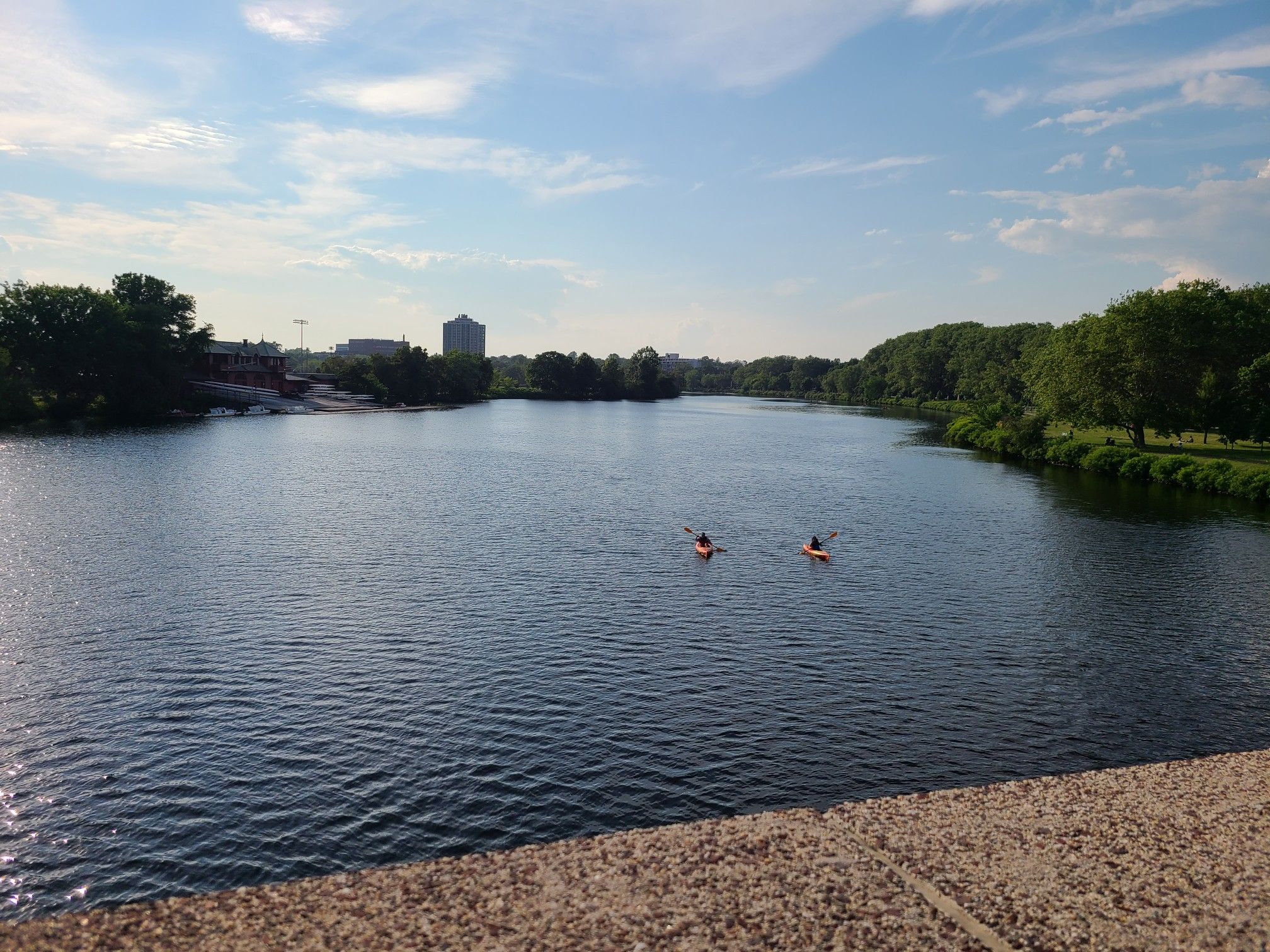 Two kayakers paddle on a wide river with distant buildings and green trees under a bright sky. - Anderson Memorial Bridge – Anderson Memorial Bridge loop from Harvard University