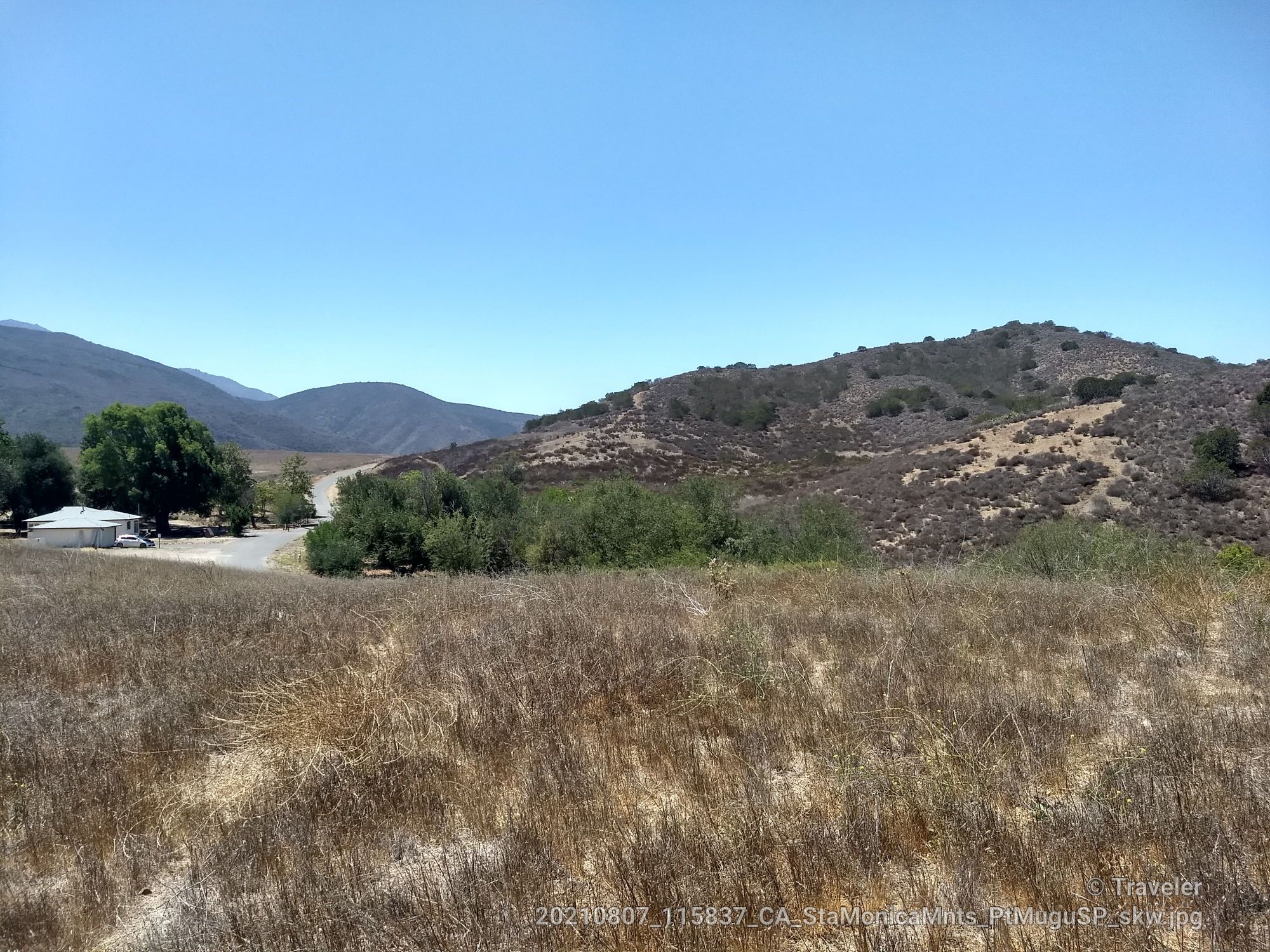 Dry grassy field with distant hills and buildings under a clear blue sky. - Rancho Potrero gravel road – Vista Del Mar Trail loop from Casa Conejo