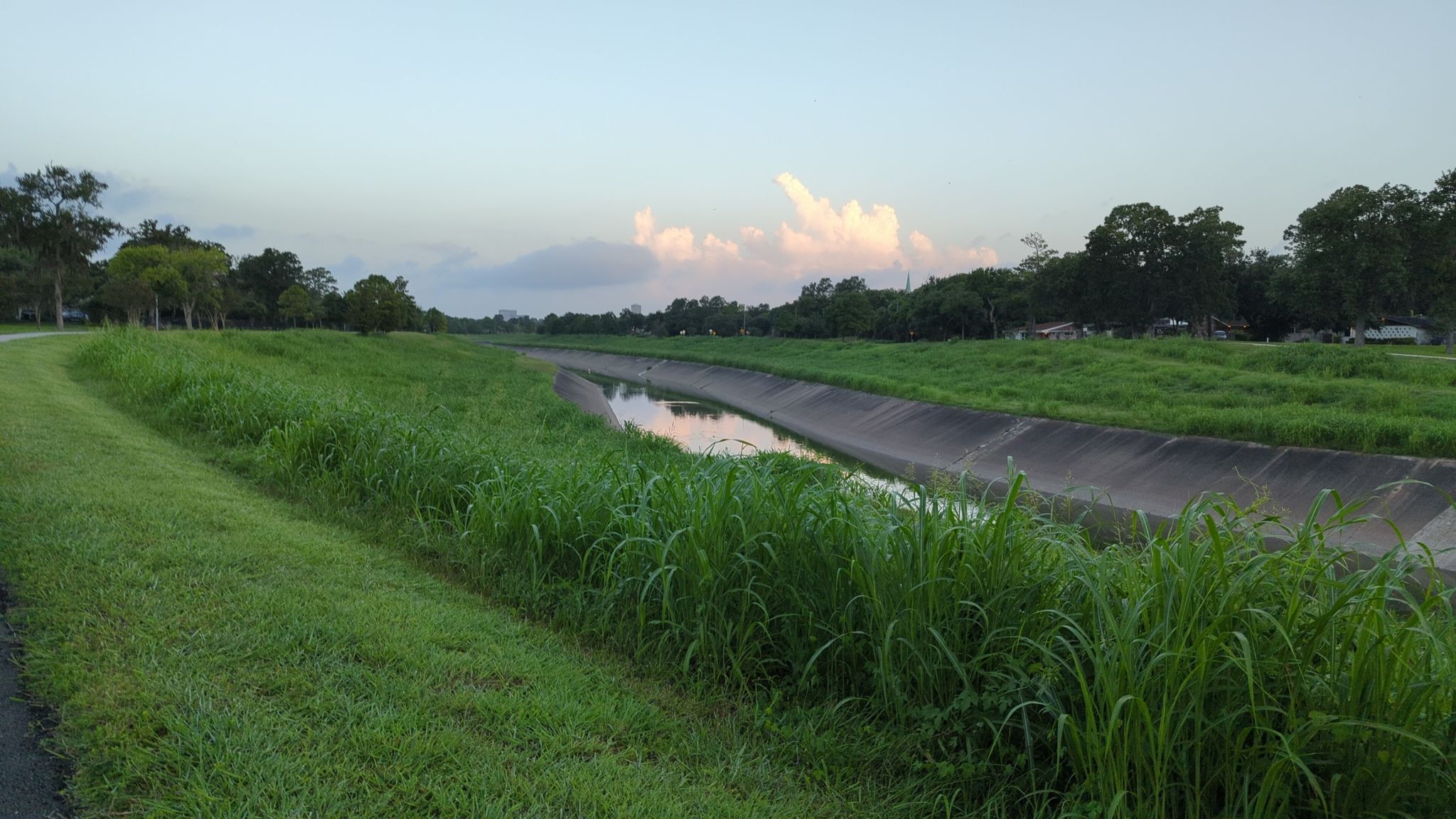 A concrete-lined canal flows between lush green grassy banks under a soft sky. - Brays Bayou Greenway – Brays Bayou Greenway loop from Central Station Capitol