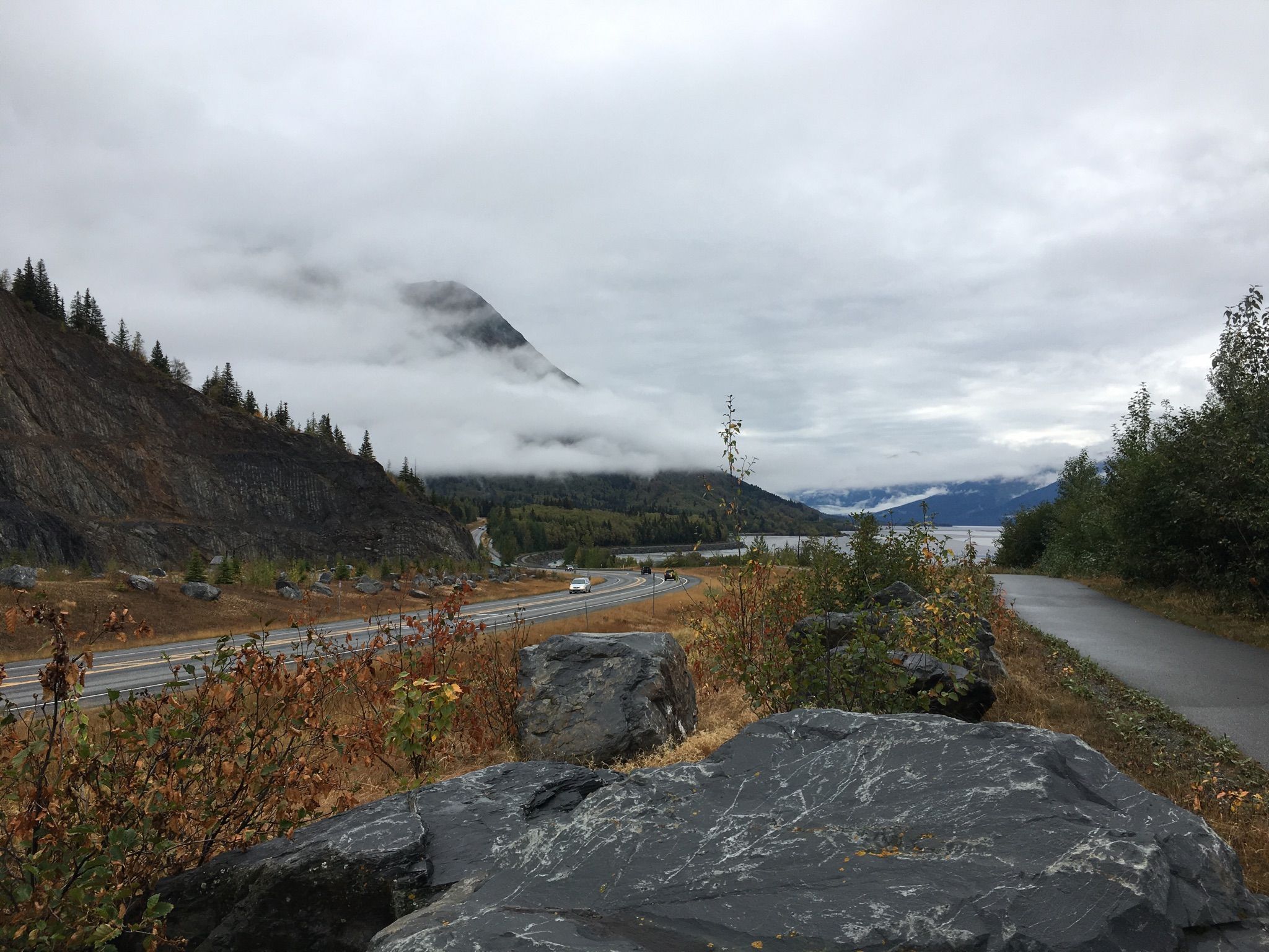 Cloudy mountain landscape with a road, water, and large rocks in the foreground. - Bird to Gird Pathway