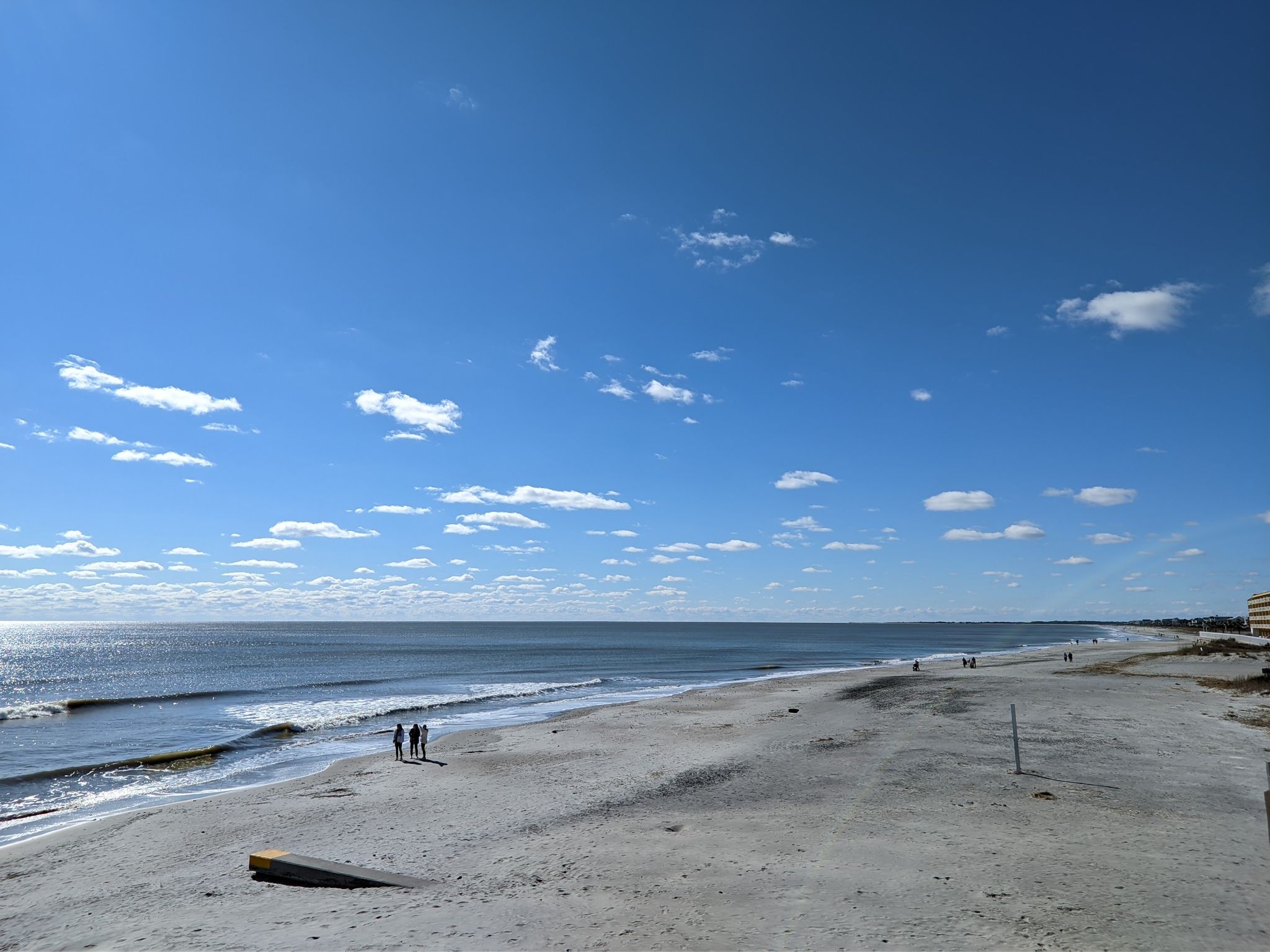 Sunny beach with ocean waves, scattered clouds, and people walking along the shore. - Folly Beach and Pier