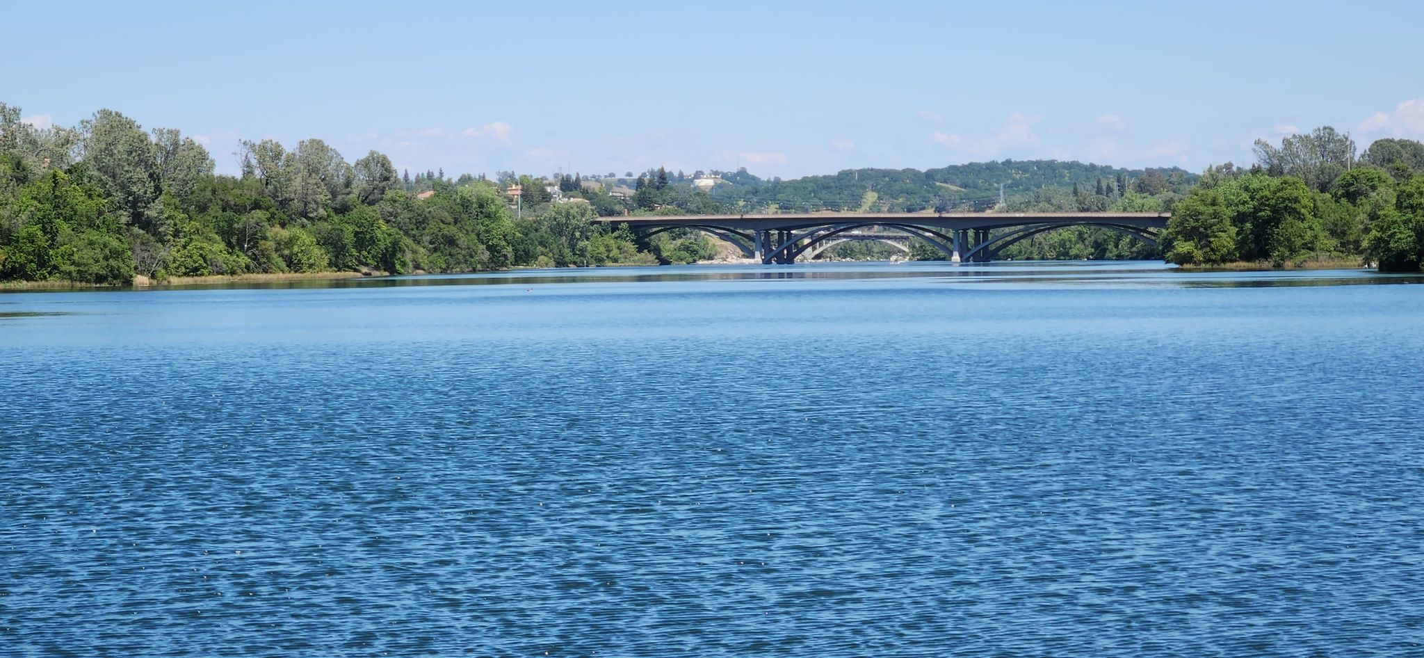 A wide blue lake with a large arched bridge in the distance under a clear sky. - Folsom Historic Truss Bridge – Folsom Railroad Museum loop from Historic Folsom