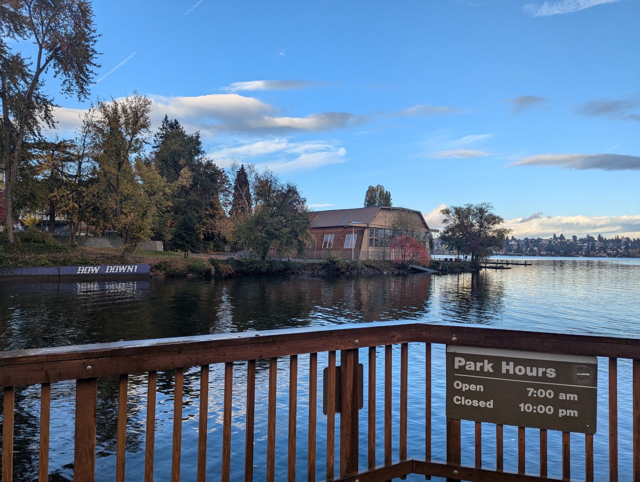 Wooden railing overlooking a lake with a building and park hours sign. - Foster Island Trail