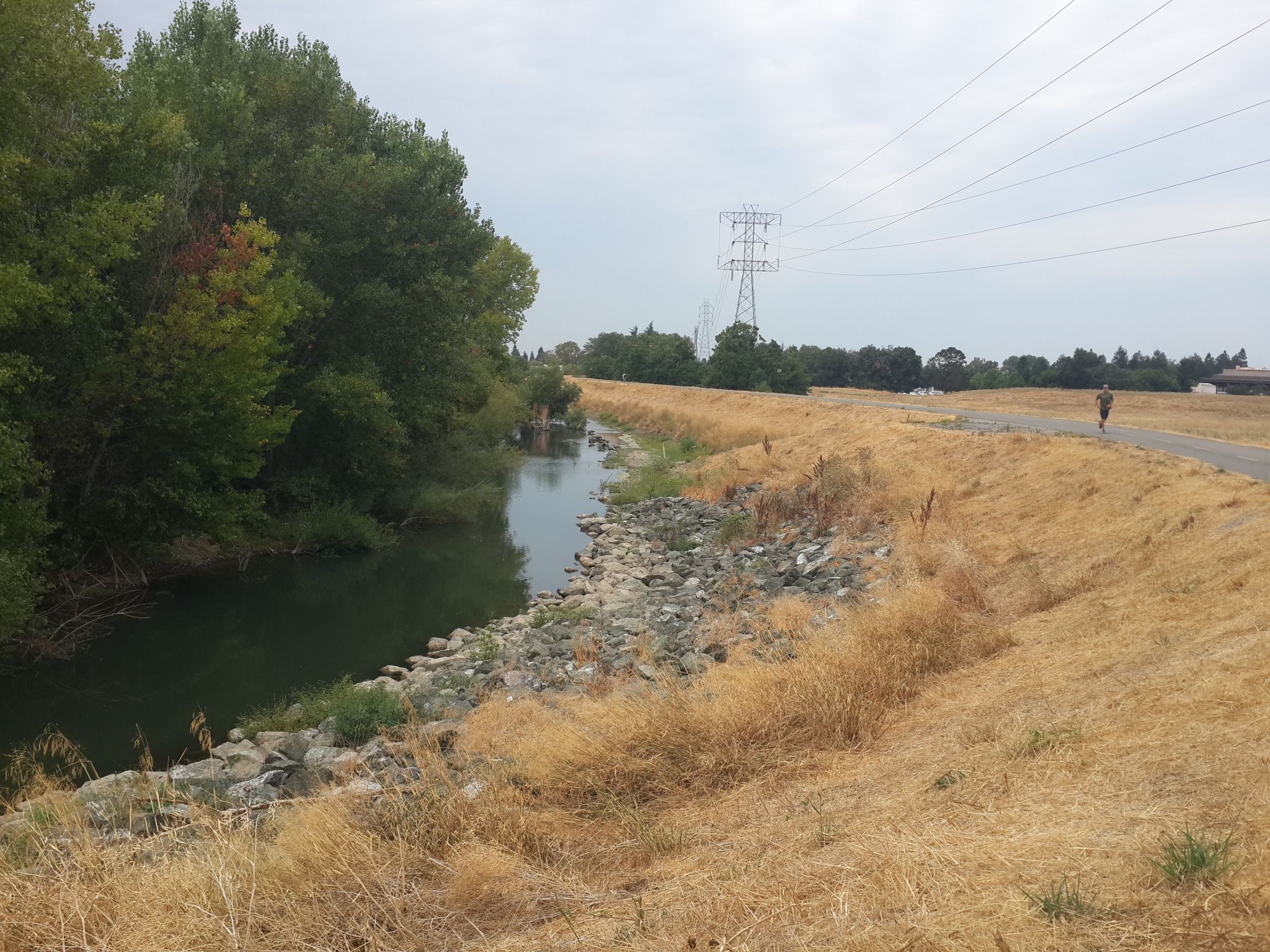 Overcast view of a river, dry path, and distant walker under power lines. - Guadalupe River Trail