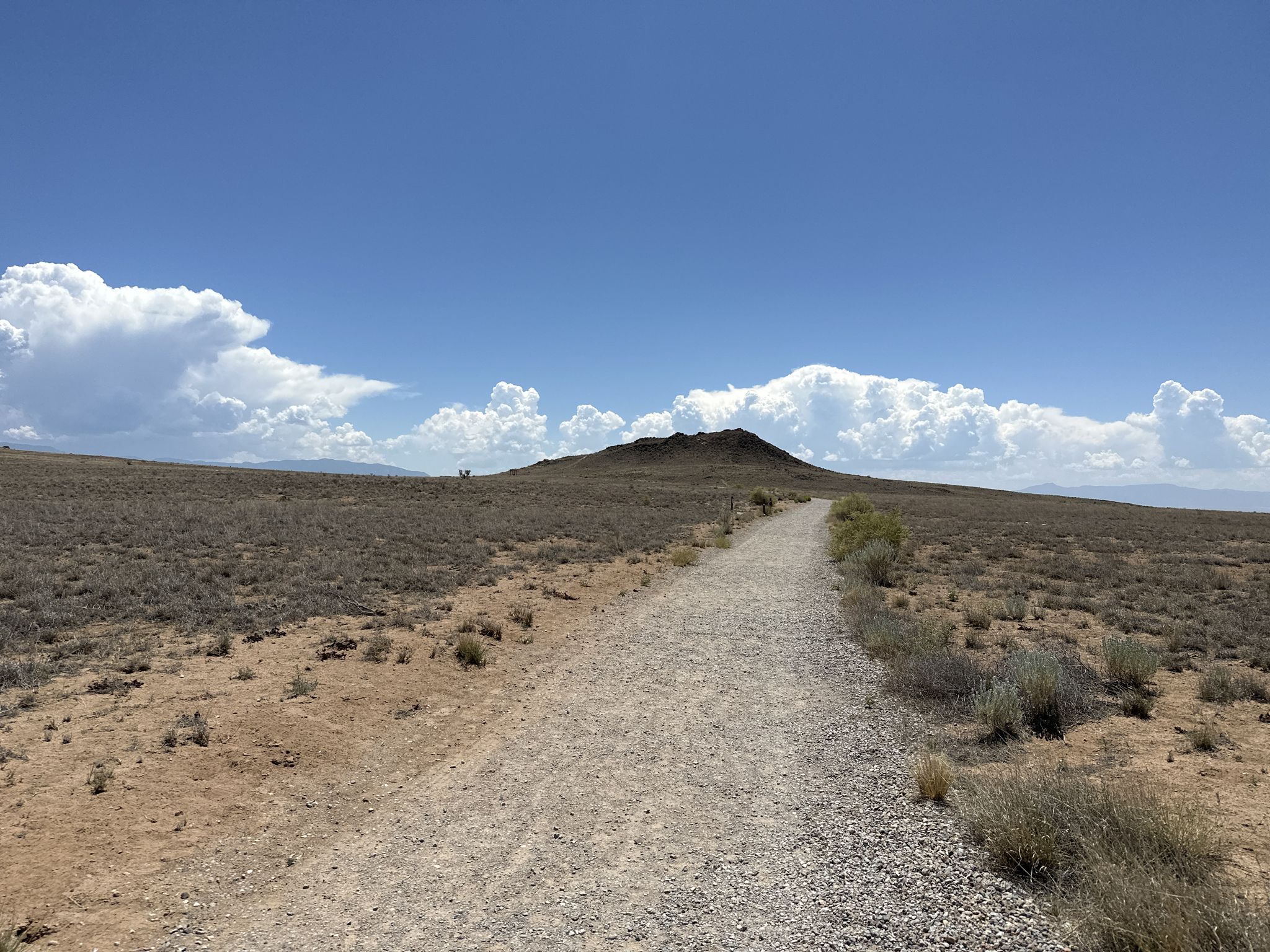Gravel path leading towards a dark hill under a bright blue sky. - Petroglyph National Monument loop