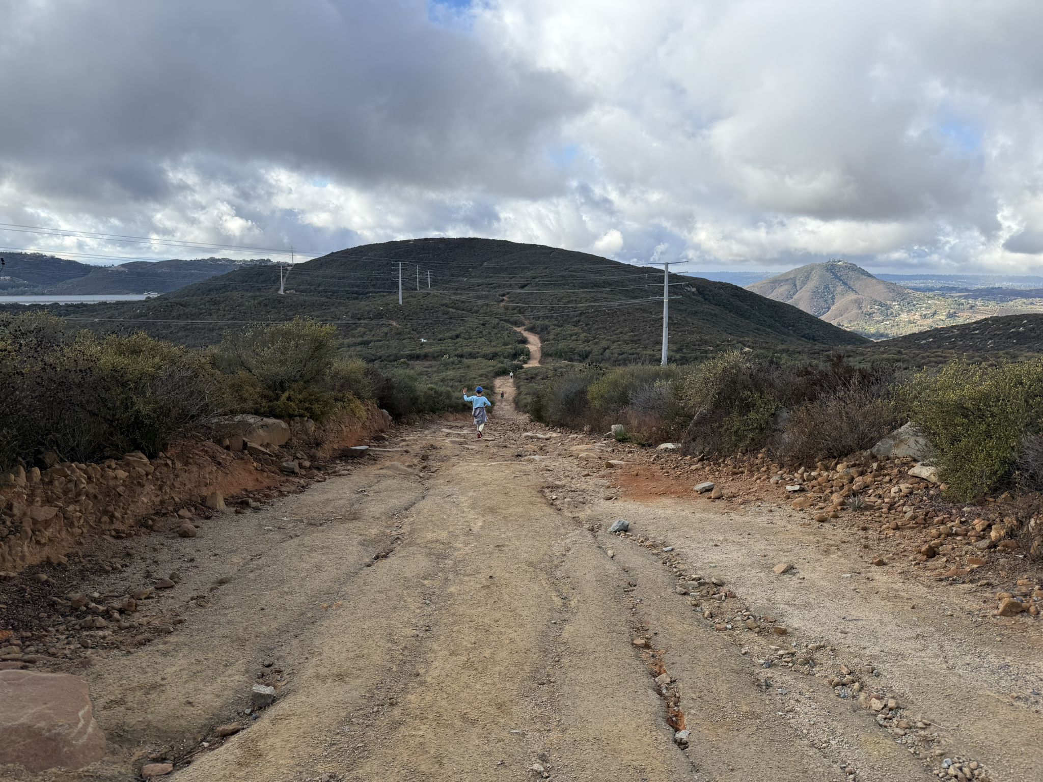 Person walks up a winding dirt road through hills under cloudy skies. - Harmony Grove Overlook – Escondido Creek loop from Harmony Grove