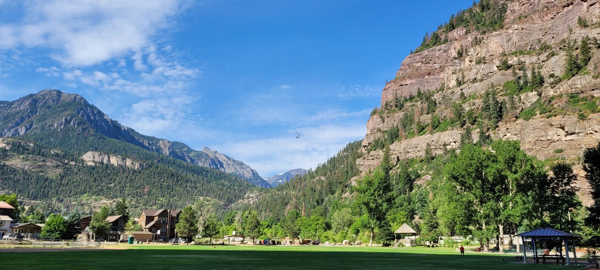 A wide grassy field with distant buildings and towering mountains under a blue sky. - Ouray – Uncompahgre River Walk Loop loop from Ouray