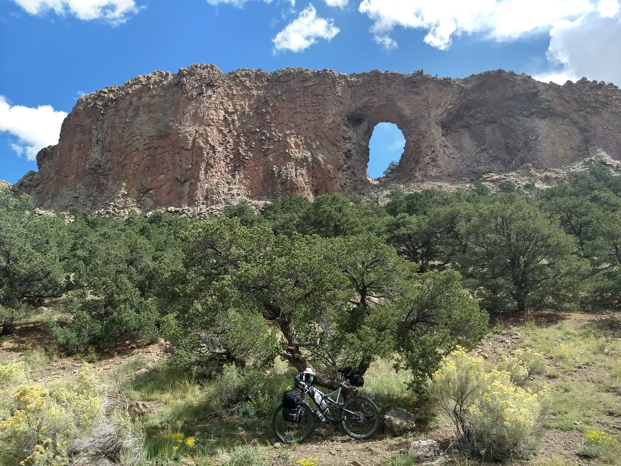 A bicycle rests under trees below a large rock arch on a sunny day. - La Garita Arch