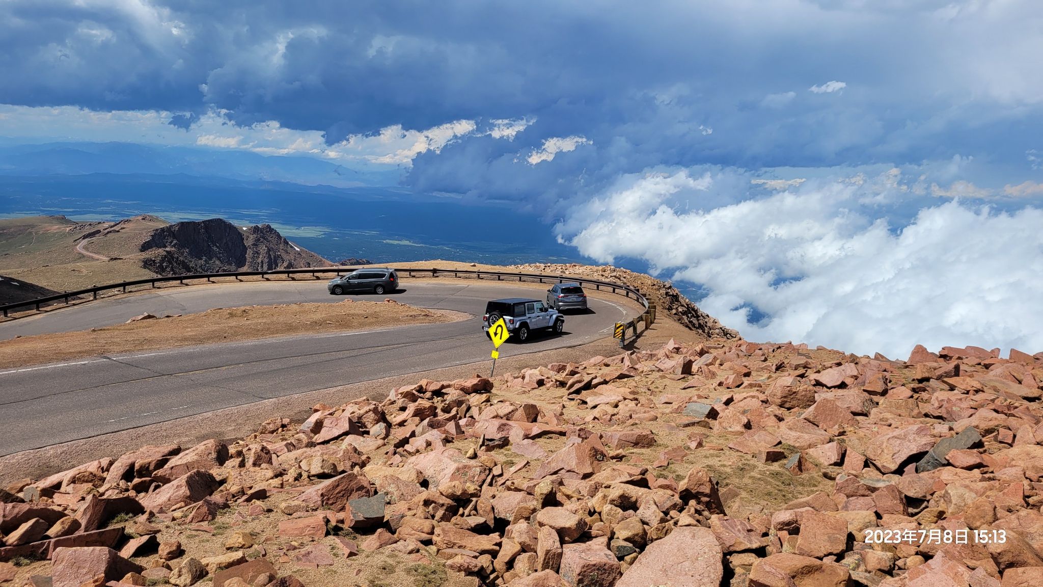 Winding mountain road with cars, rocky terrain, and clouds below. - Pikes Peak from Devils Playground