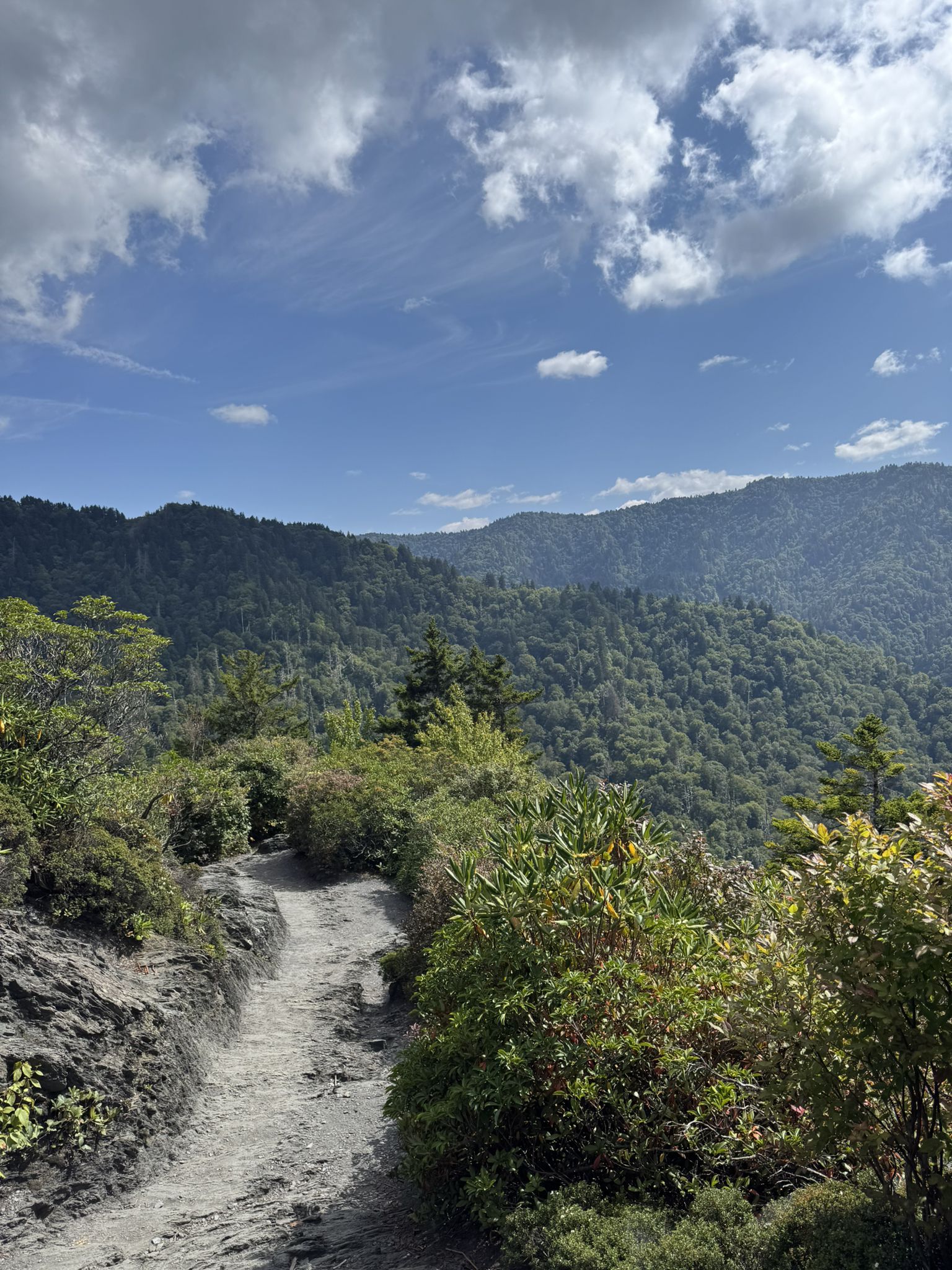A narrow dirt path winds along a mountain ridge with distant green peaks. - Alum Cave Trail