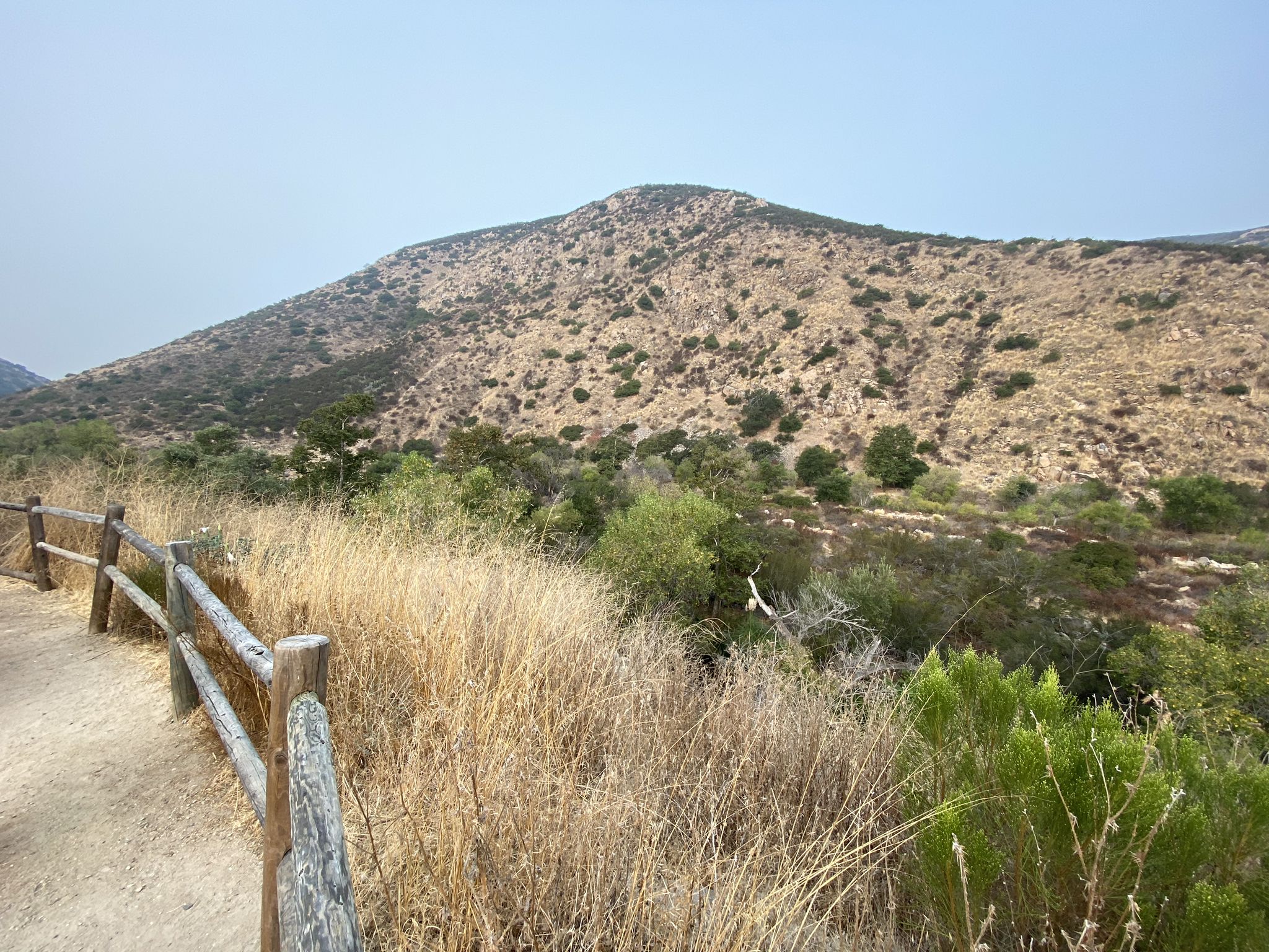 Dirt path with wooden fence beside a dry, brushy hillside under clear sky. - Old Mission Dam – Mission Trails Regional Park loop from Gillespie Field